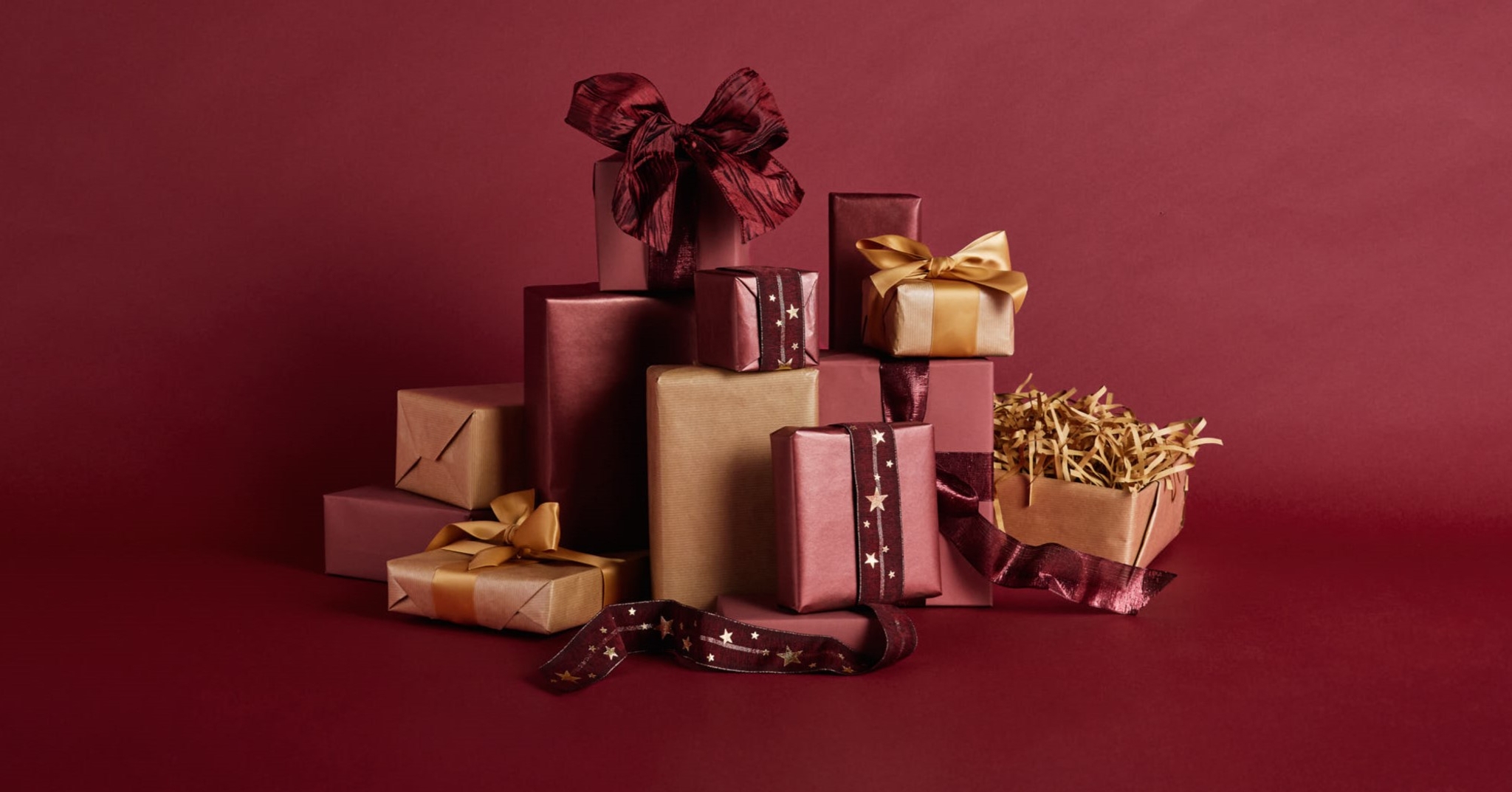 A pile of gift boxes wrapped in shades of red, burgundy, and kraft, decorated with gold and star ribbons, against a crimson background.
