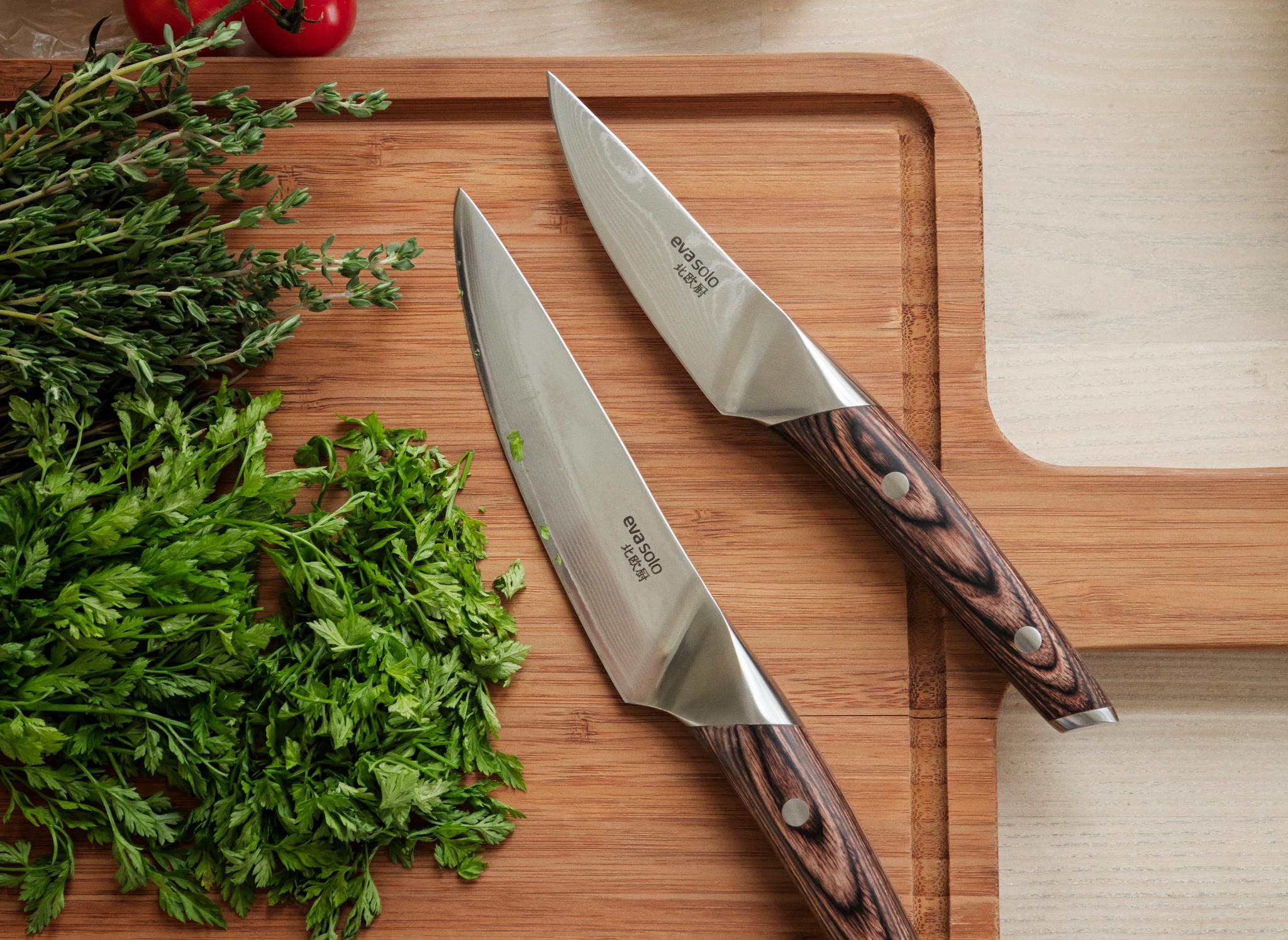 Kaksi veistä ja tuoreita yrttejä puisella leikkuulaudalla, taustalla tomaatteja ja valkosipulia muovipusseissa.