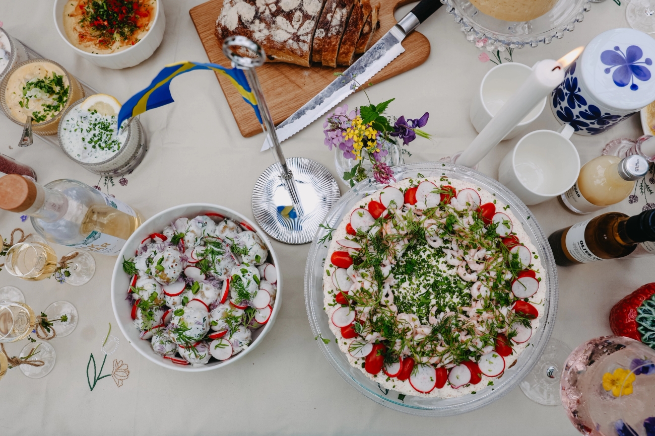 Somrig svensk buffé med smörgåstårta, sill, bröd och dryck uppdukat på festligt dukat bord.