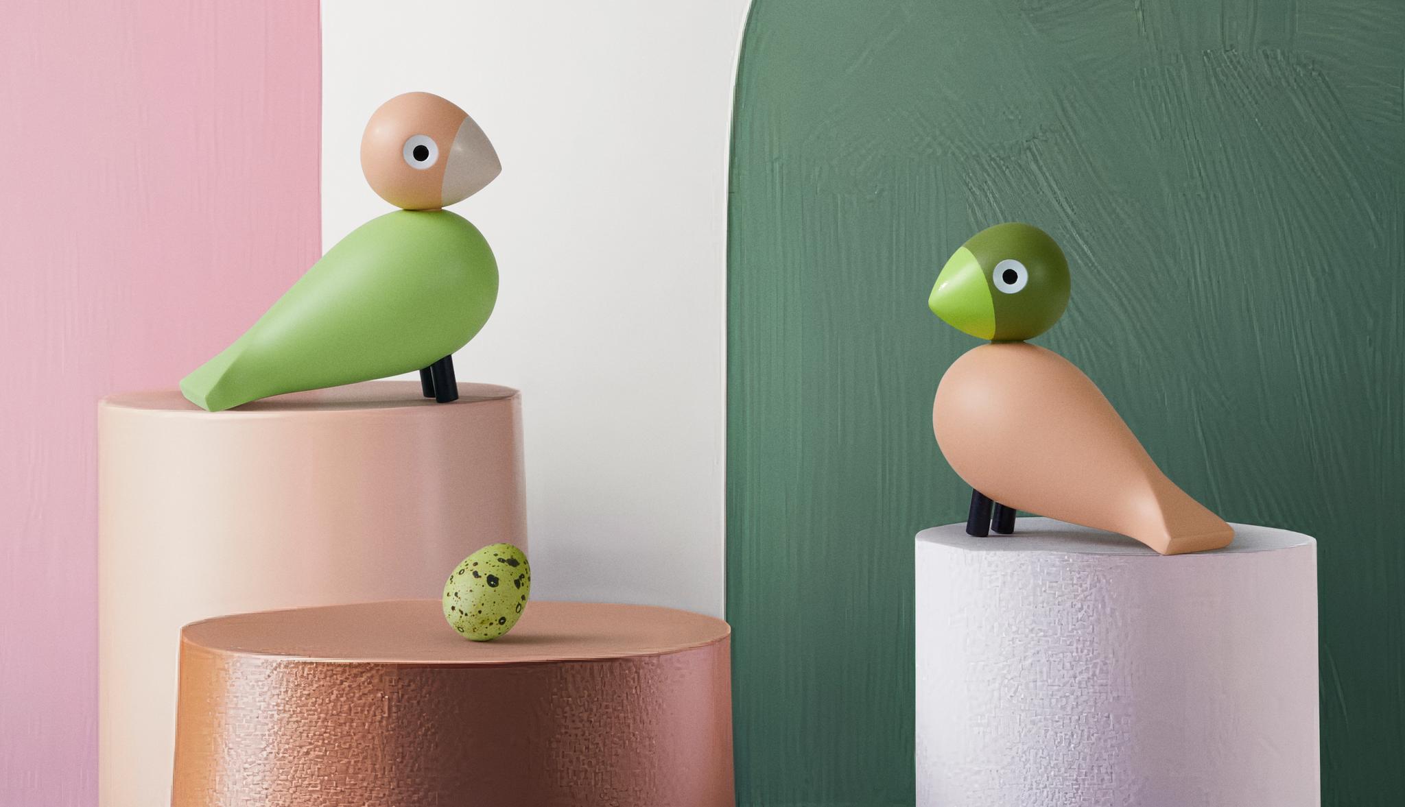 Värikkäät puiset design-linnut ja pilkullinen muna on aseteltu pastellisävyisille jalustoille graafisen, maalatun taustan eteen.
