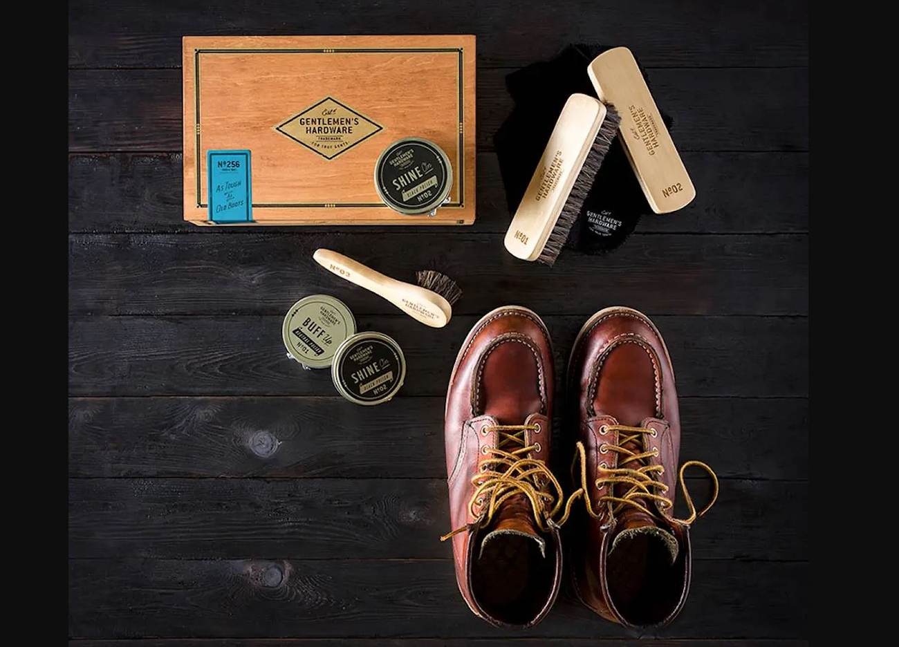 A shoe care kit with polishes and brushes arranged next to a pair of brown leather boots on a dark wooden surface.