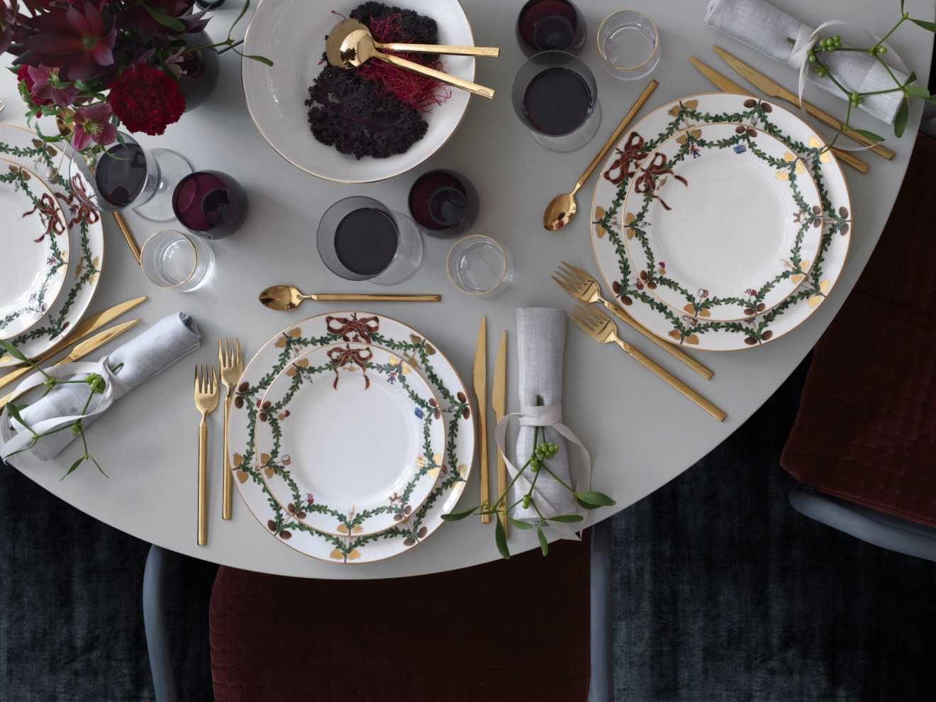 Ylhäältä katsottuna elegantti ruokapöytä, jossa on valkoisia juhla-aiheisia lautasia, kultaisia ​​aterimia, viiniä ja vihreillä sidottuja lautasliinoja.