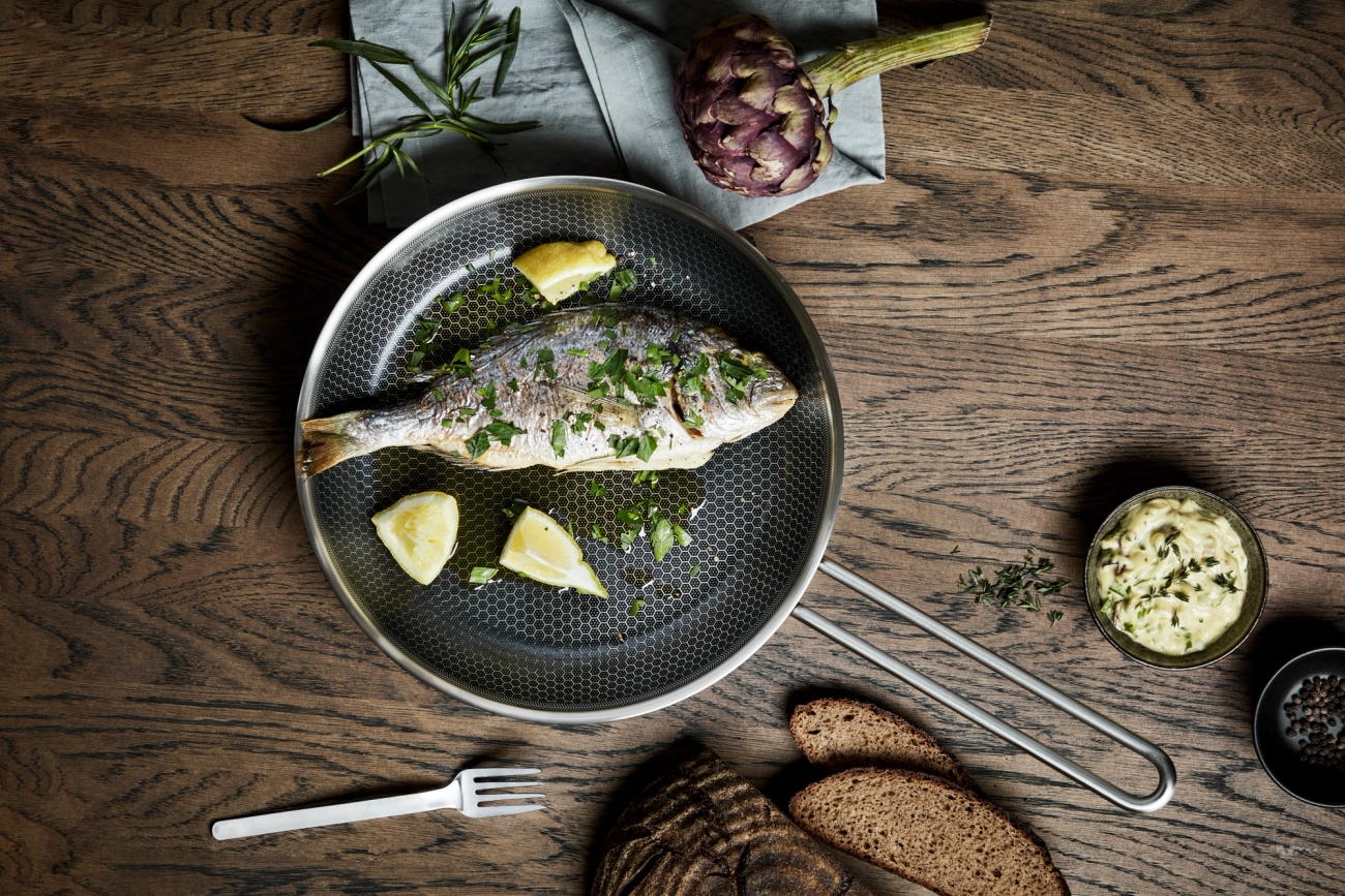 Whole cooked fish with lemon and herbs in a frying pan on a rustic wooden table.