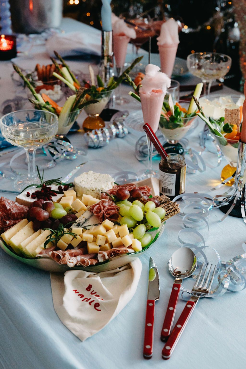 Ett festbord rymmer en stor charkuteribricka, champagneglas, rosa sockervadd och crudités på en blå duk.
