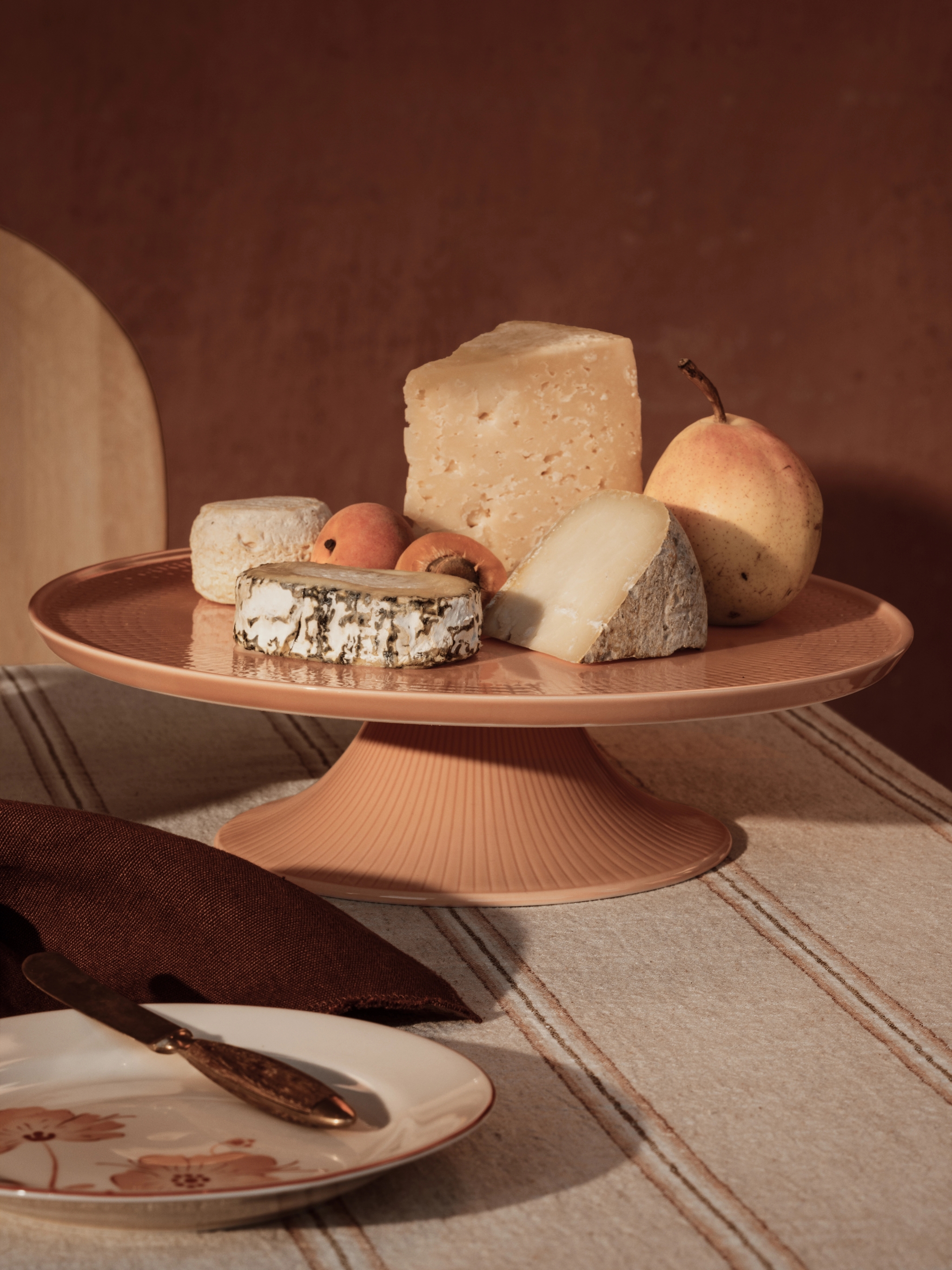 Et kakefat med ulike oster, en pære og aprikoser på en stripete duk, med en tallerken og kniv i forgrunnen.