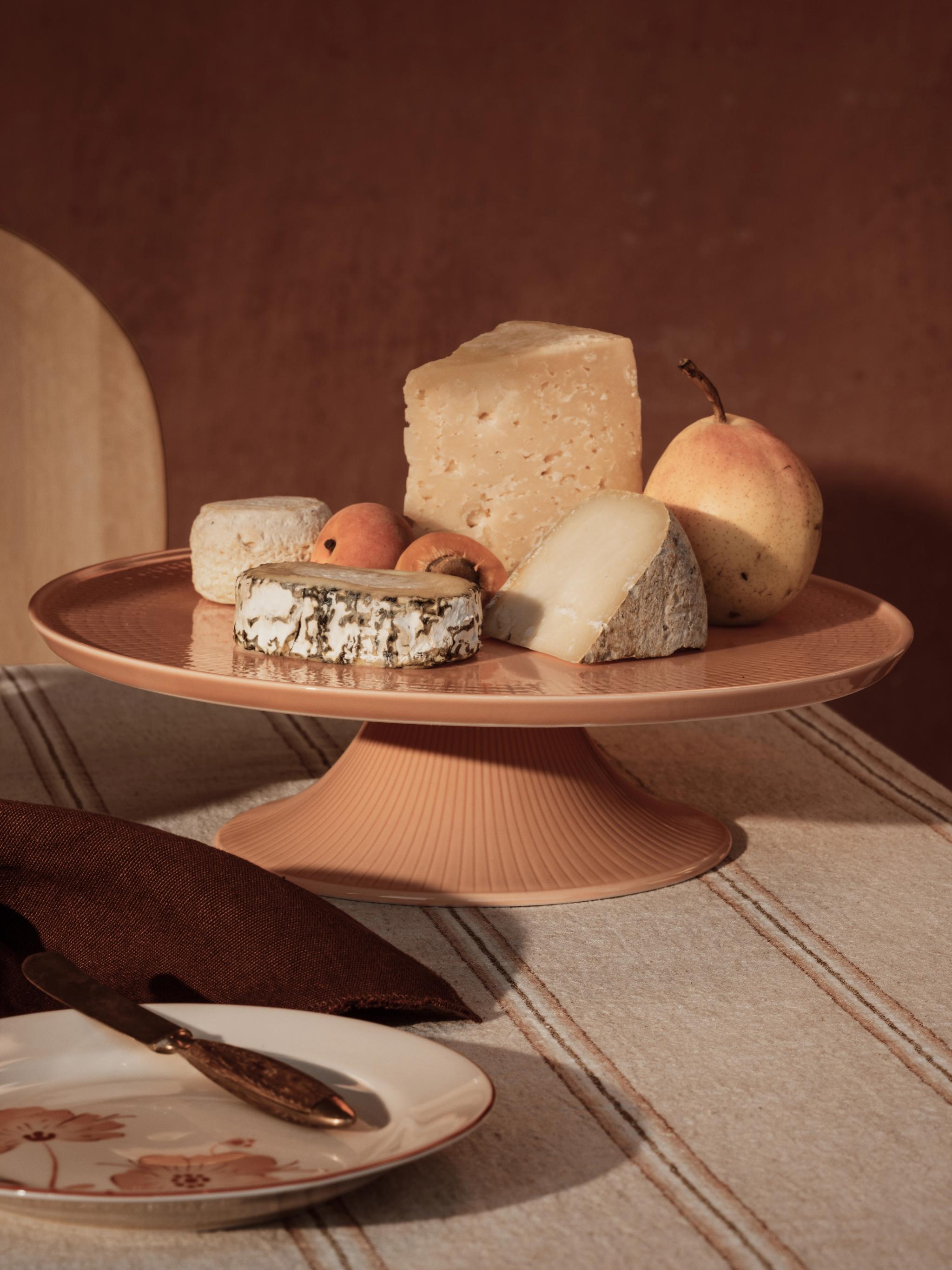 Et kakefat med ulike oster, en pære og aprikoser på en stripete duk, med en tallerken og kniv i forgrunnen.
