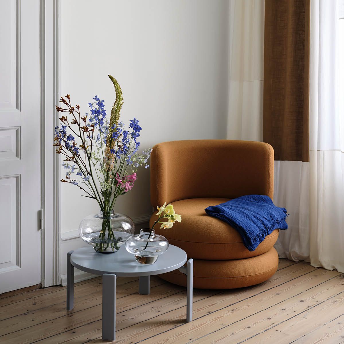  En orange fåtölj med en blå filt bredvid ett runt blått bord med två vaser med färgglada blommor