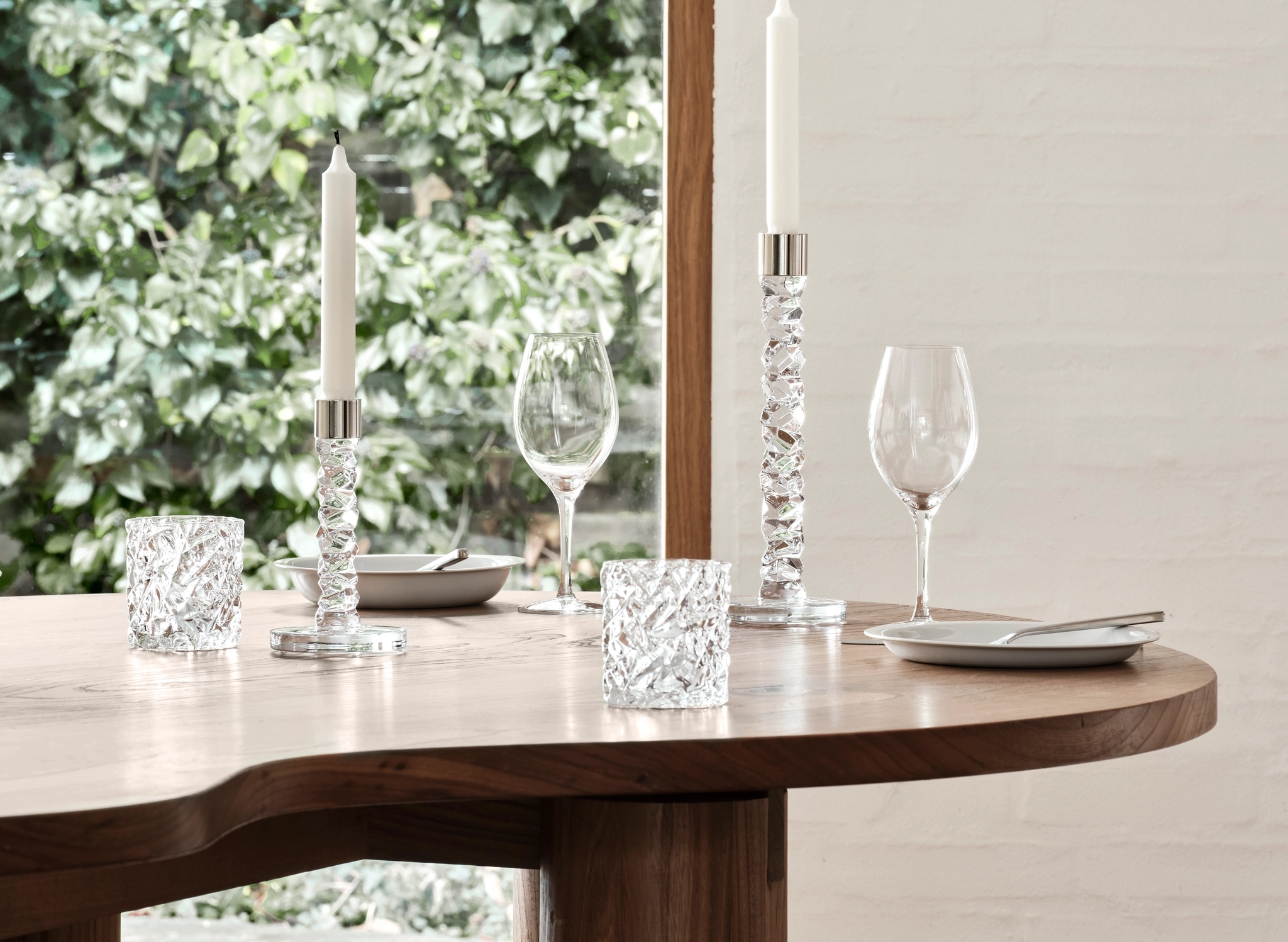 Vinglas, ljusstakar och ljuslyktor från Orrefors Carat på ett träbord med en grönskande trädgård i bakgrunden.