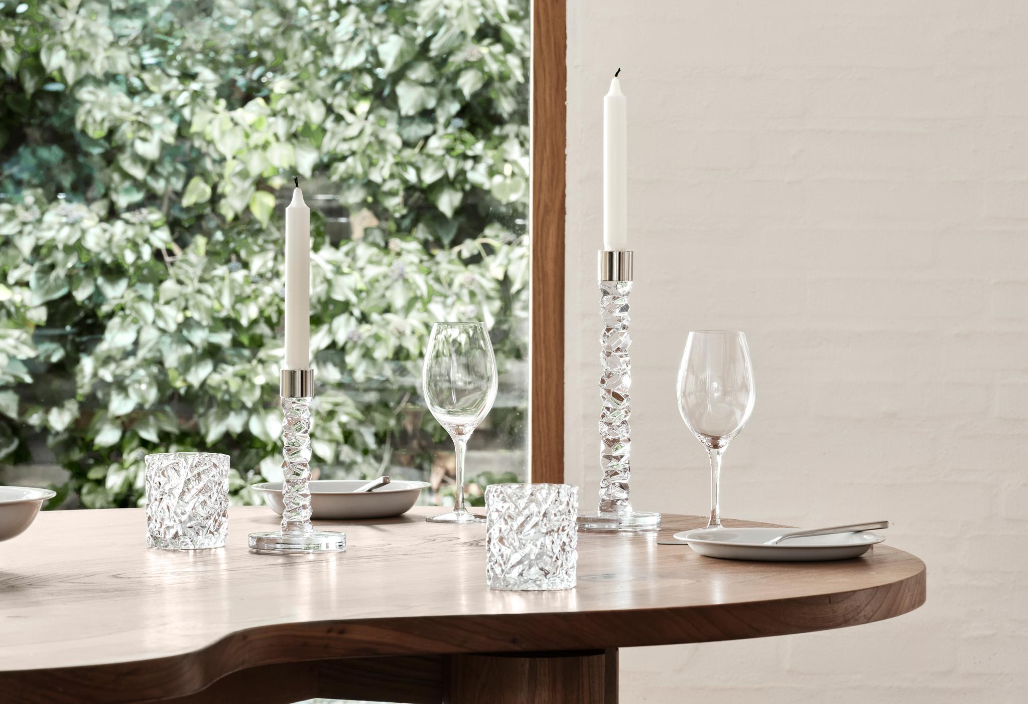 Vinglas, ljusstakar och ljuslyktor från Orrefors Carat på ett träbord med en grönskande trädgård i bakgrunden.