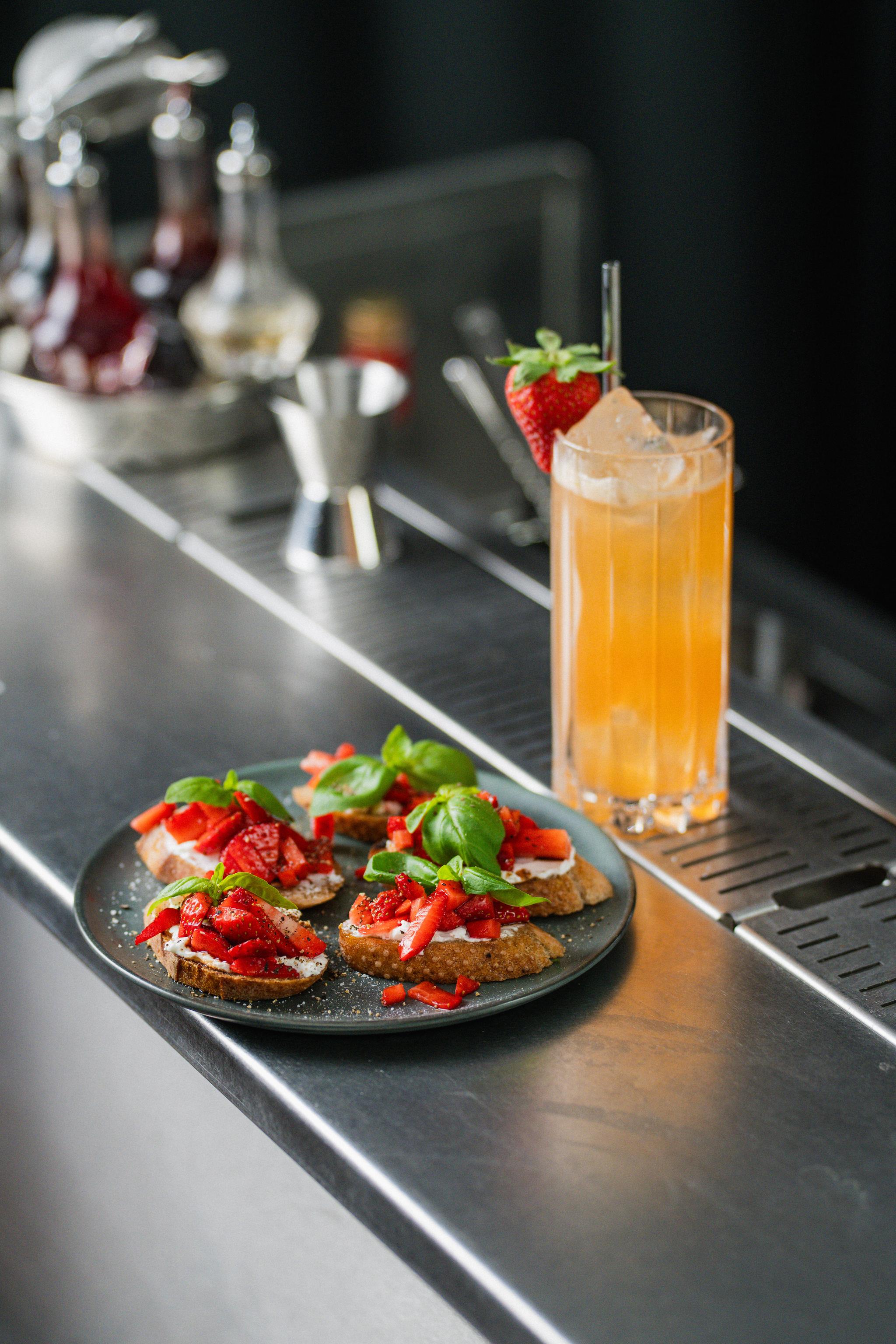 En tallrik med crostini toppade med jordgubbar och basilika står på en bardisk bredvid ett högt glas orange drink med is och jordgubbsdekor.