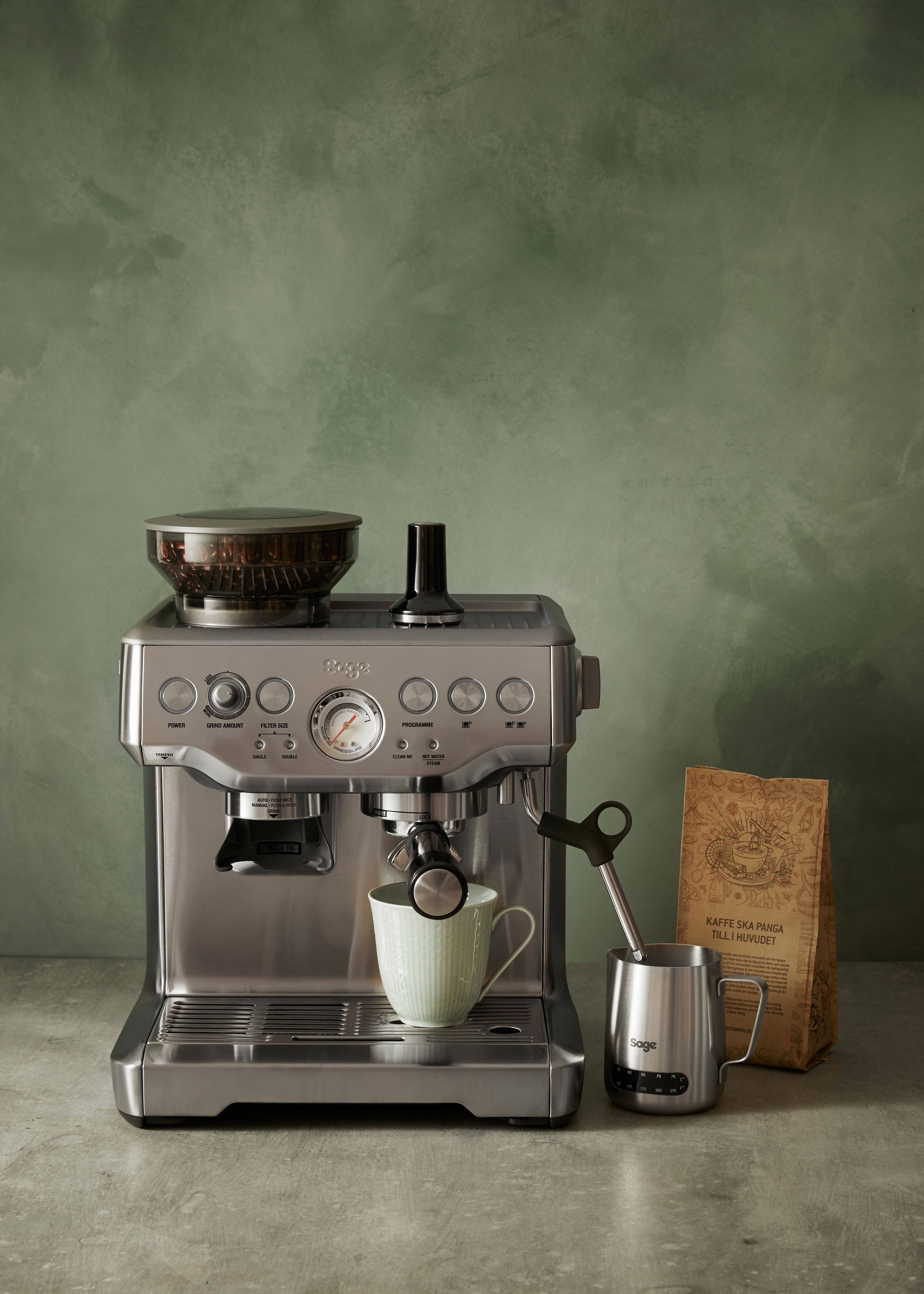 Tyylikäs Sage-espressokone keittiötasolla kahvipussin ja maitokannun kanssa vihreää seinää vasten.
