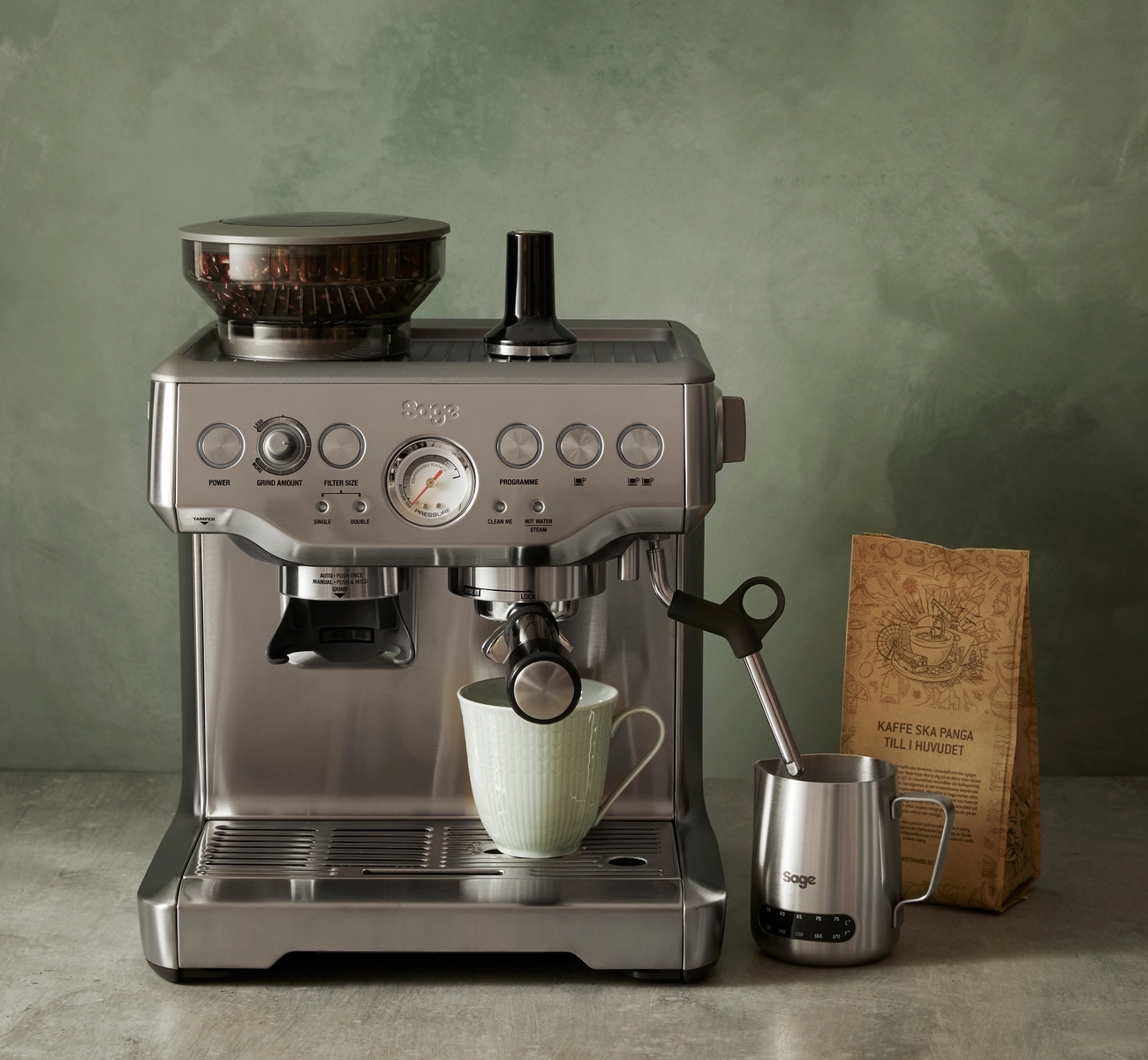 Tyylikäs Sage-espressokone keittiötasolla kahvipussin ja maitokannun kanssa vihreää seinää vasten.