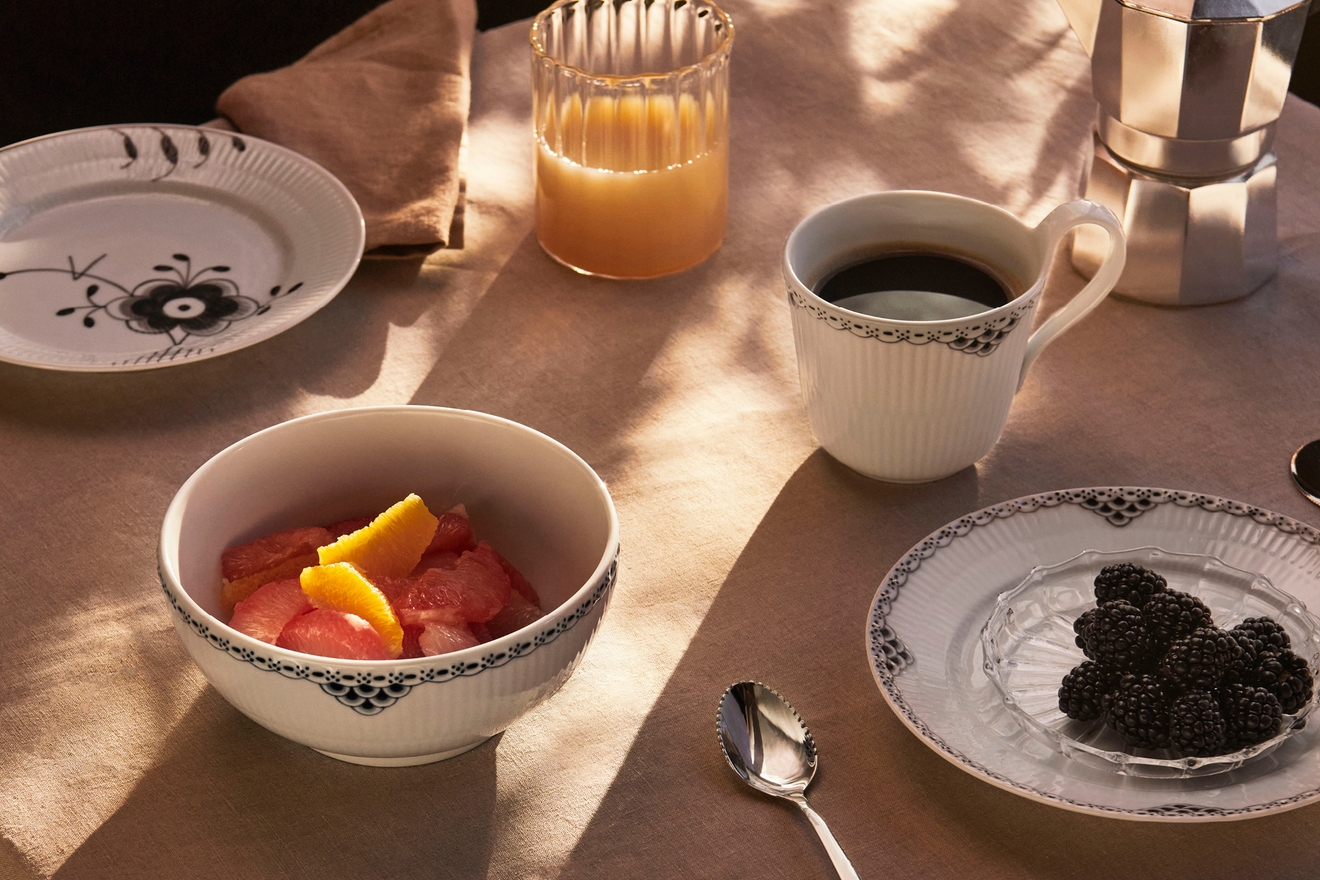 Frukostbjudning med kaffe, apelsinjuice, grapefrukt och björnbär serverade i vitmönstrade fat på ett soligt bord.