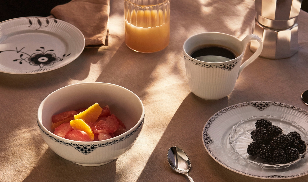 Frukostbjudning med kaffe, apelsinjuice, grapefrukt och björnbär serverade i vitmönstrade fat på ett soligt bord.