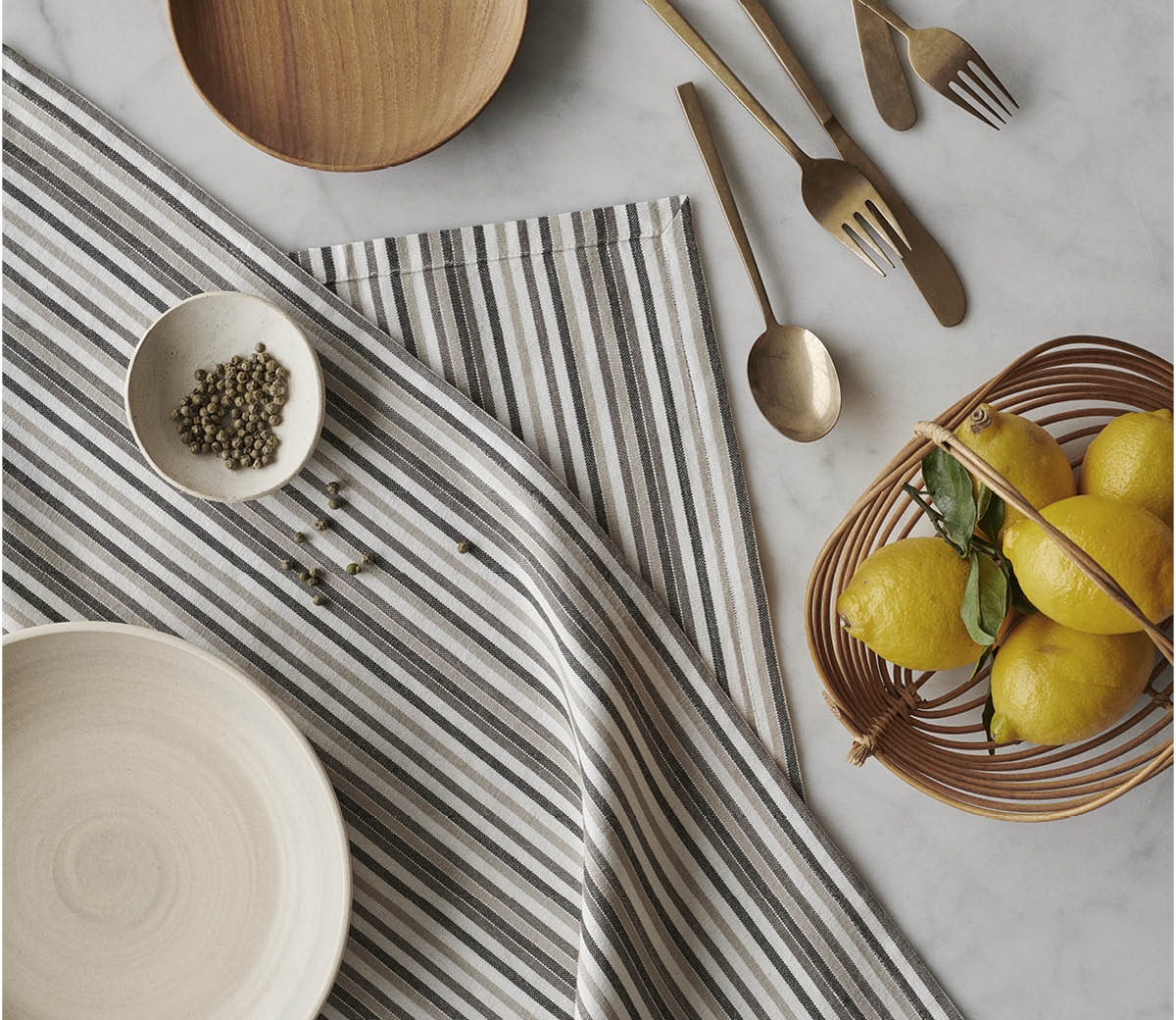 Kuva pöydästä, jossa raidallinen By Mogensen liina, puisia ja keraamisia astioita, kullanväriset aterimet, vihreitä pippureita ja sitruunoita korissa.