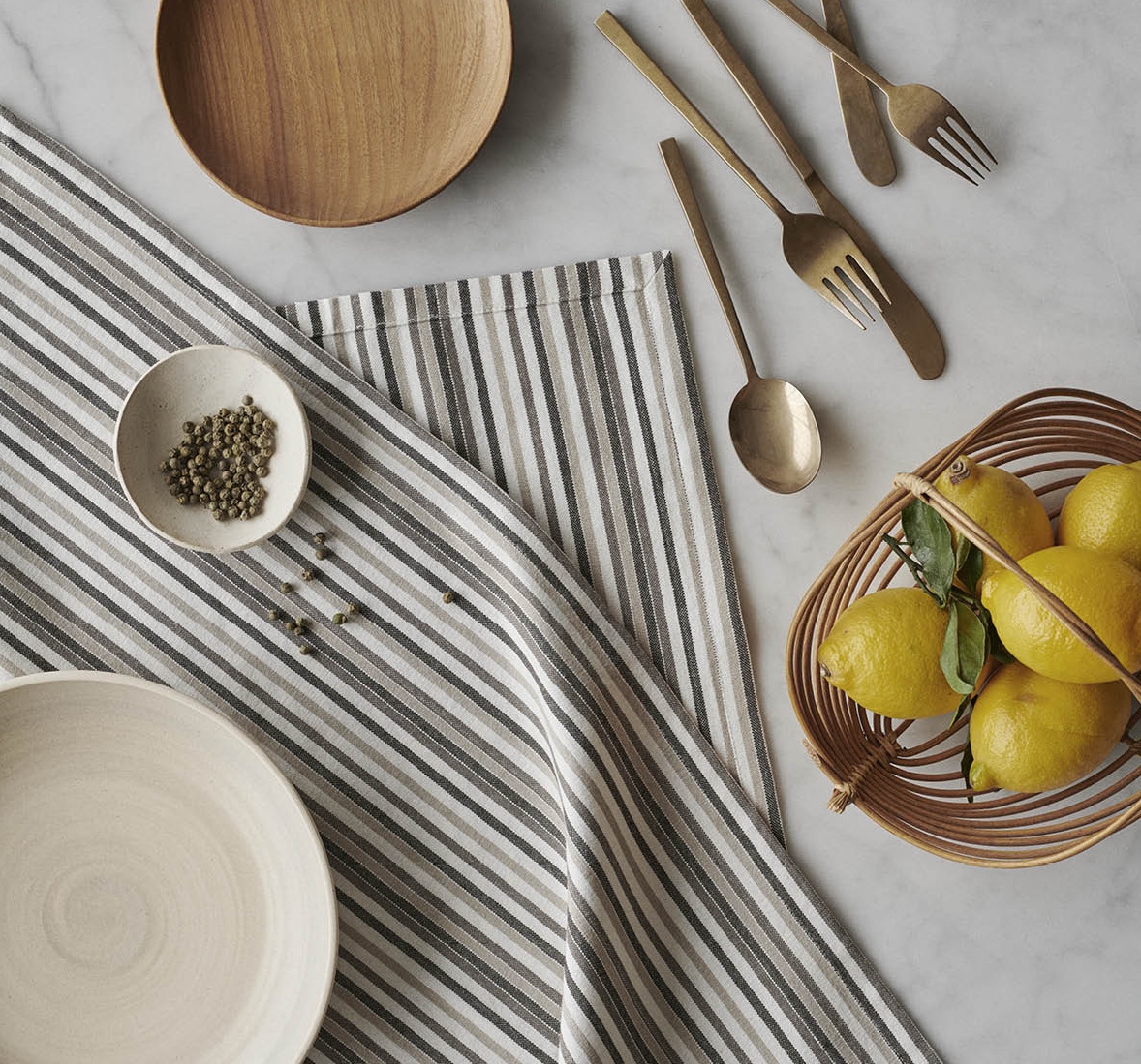 Kuva pöydästä, jossa raidallinen By Mogensen liina, puisia ja keraamisia astioita, kullanväriset aterimet, vihreitä pippureita ja sitruunoita korissa.