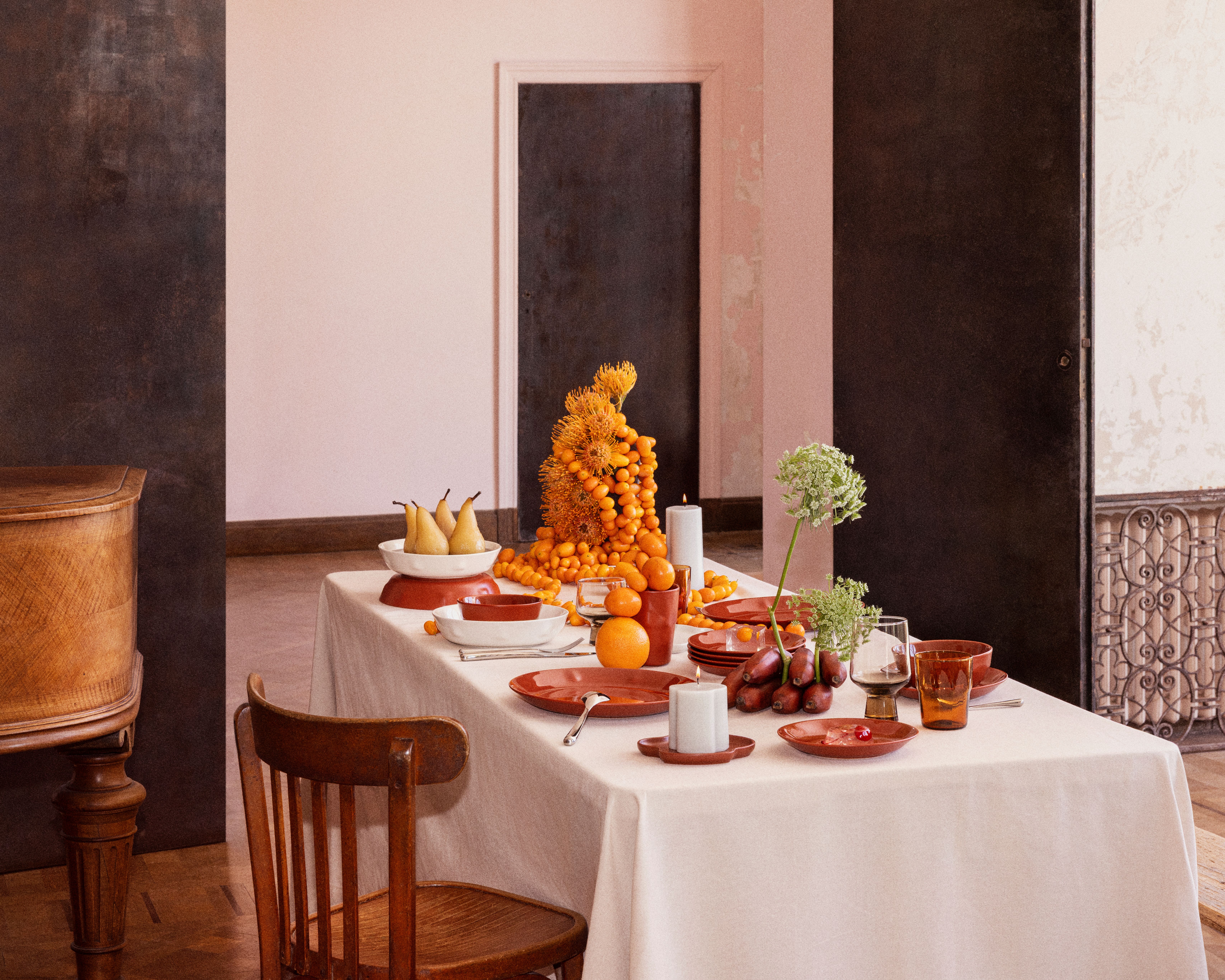 Ruokapöytä, jossa on terrakotta-astioita, valkoinen pöytäliina ja eloisa oranssien hedelmien ja keltaisten kukkien muodostama pöytäkoriste.