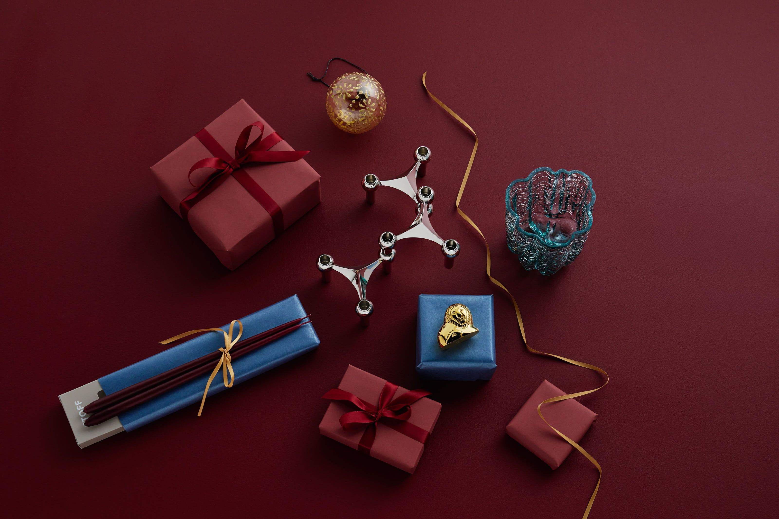 Festligt arrangemang av presenter, en silverljusstake, en guldprydnad och dekorativa föremål på en djupröd yta.