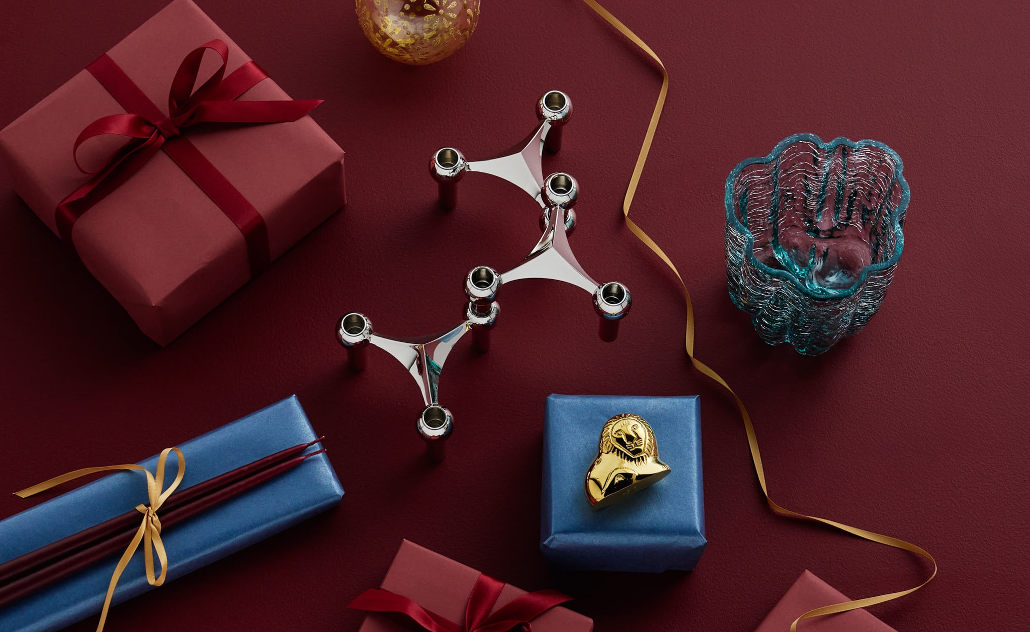 Festive gifts and decorations, including a silver candelabra and blue glass bowl, on a red background.