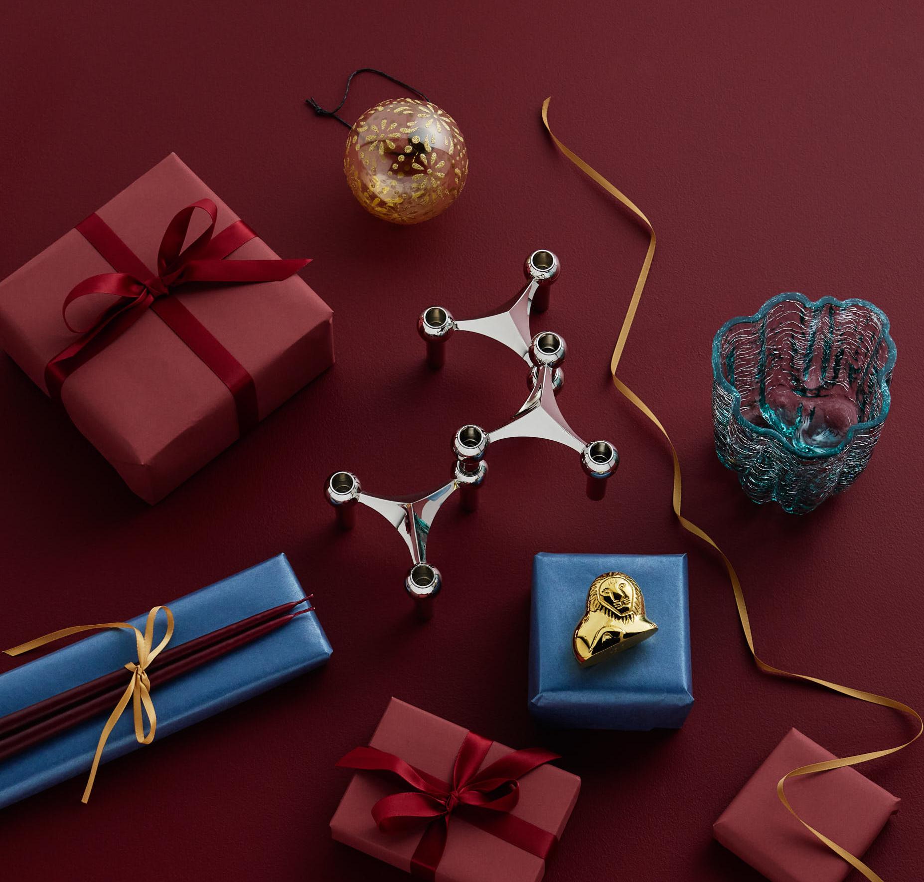 Various wrapped gifts, a silver candelabra, blue glass bowl, chopsticks, and a gold ornament arranged on a red surface with a gold ribbon.