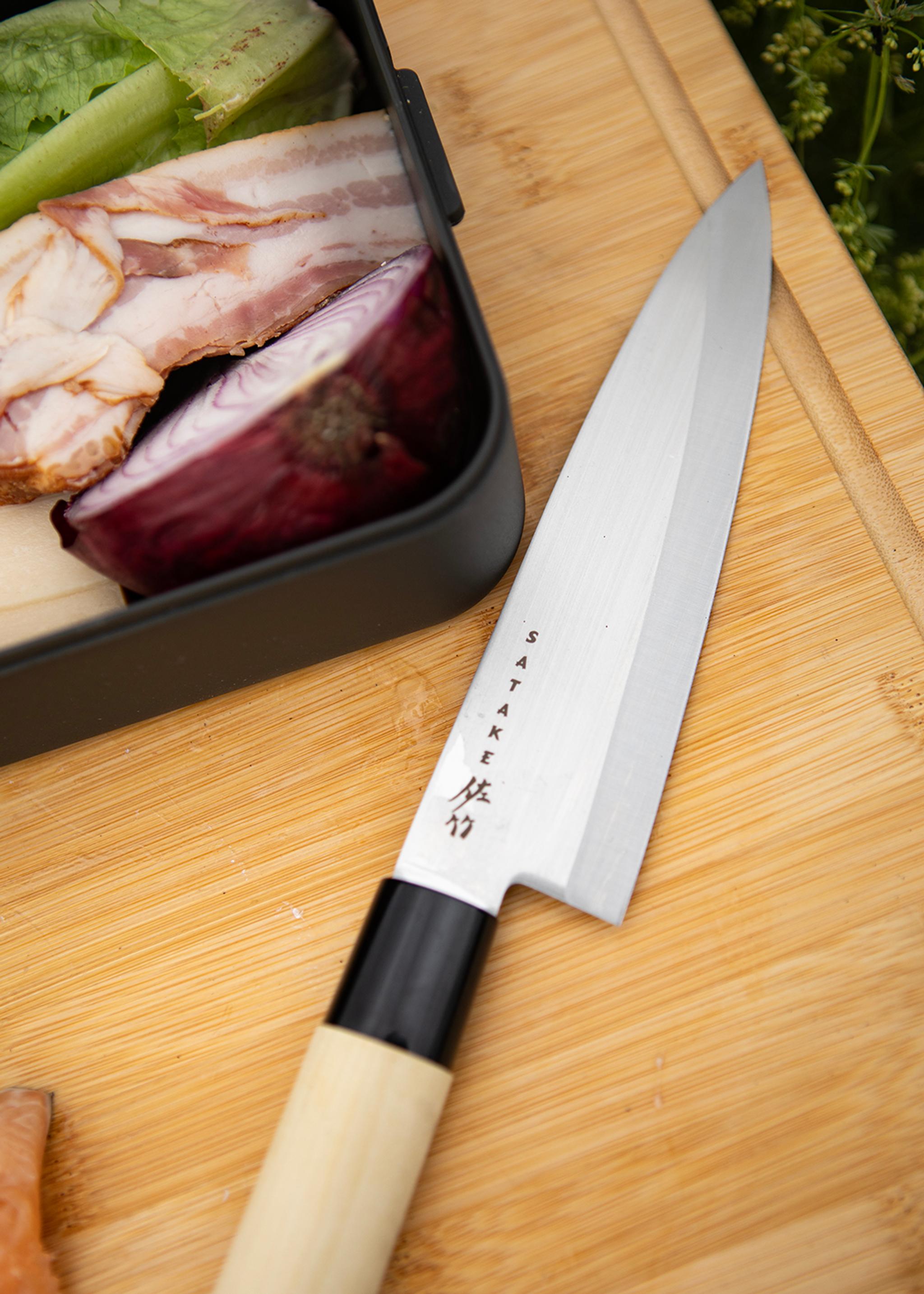 Japansk Satake-kockkniv ligger på en skärbräda bredvid grönsaker, bacon och rödlök.