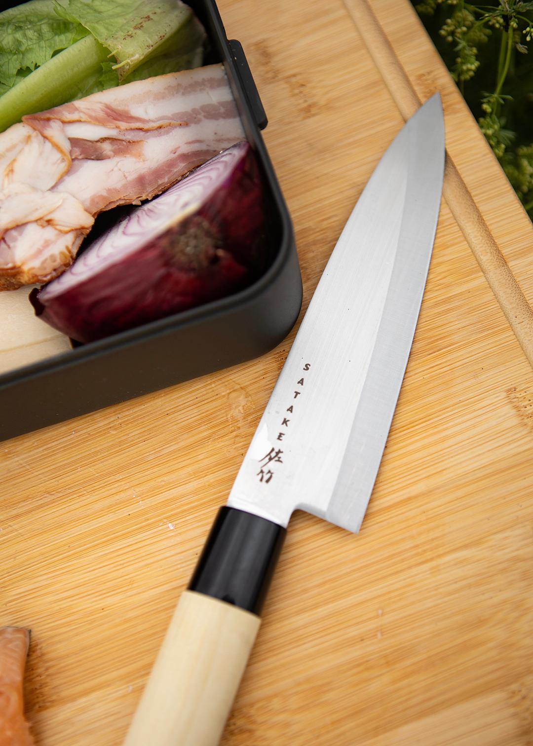 Japansk Satake-kockkniv ligger på en skärbräda bredvid grönsaker, bacon och rödlök.