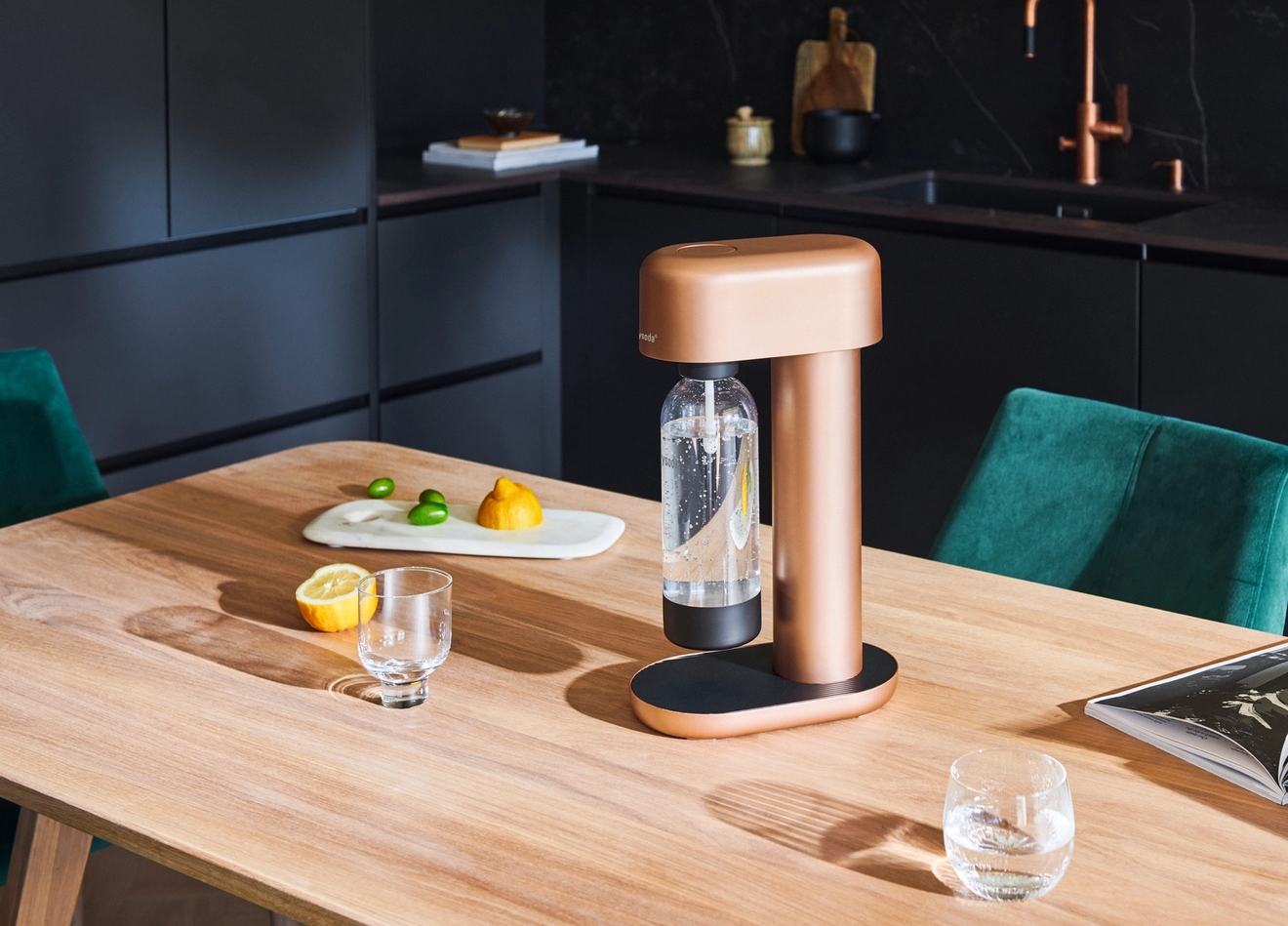 Ruusukullanvärinen hiilihapotuslaite puisella pöydällä sitruunoiden, vesilasien ja avoimen kirjan kanssa modernissa, tummasävyisessä keittiössä.