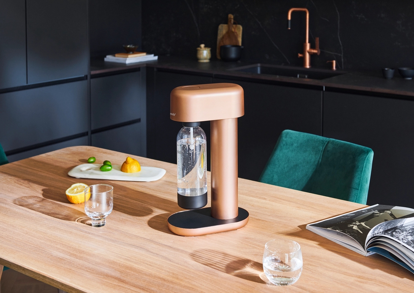 A copper soda maker with a bottle of sparkling water, lemons, limes, and glasses on a wooden table in a modern kitchen.