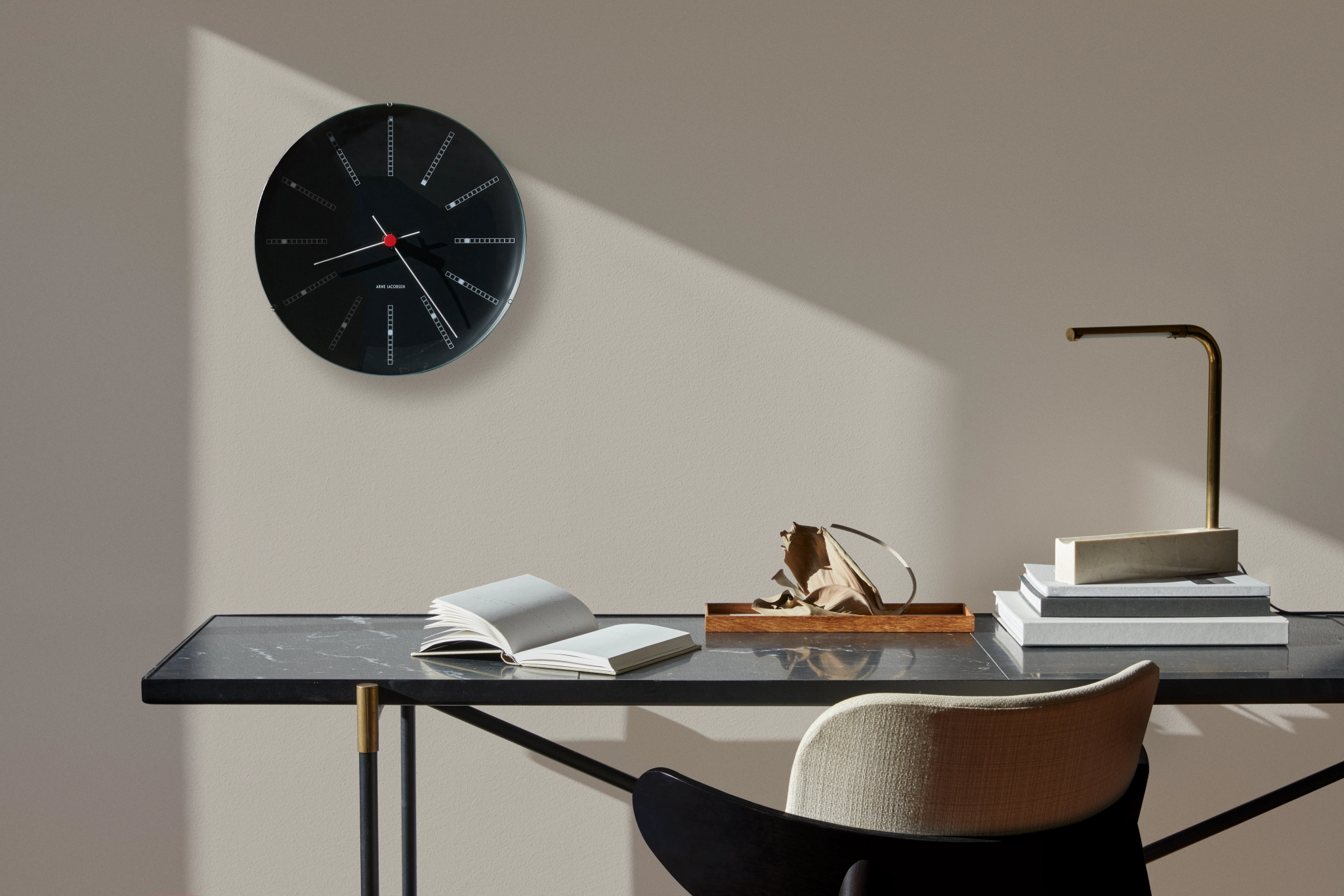 Musta Arne Jacobsen seinäkello roikkuu tumman työpöydän yläpuolella, jonka päällä on kirja, messinkilamppu ja koriste-esineitä auringonvalossa