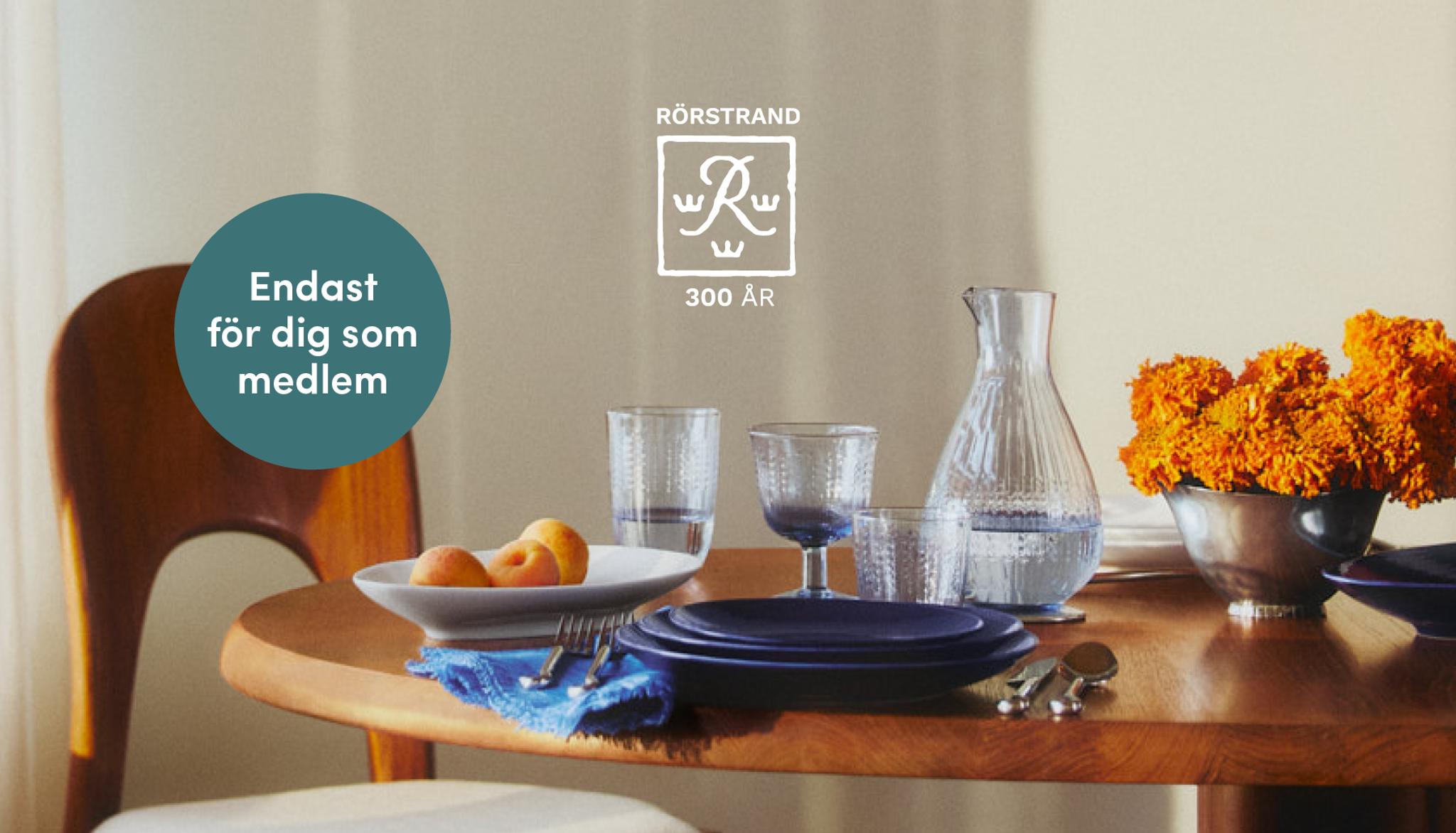 Ett matbord med mörkblå tallrikar, texturerade glas, en vattenkaraff, orange blommor och aprikoser. Text: RÖRSTRAND 300 ÅR och "Endast för dig som medlem".