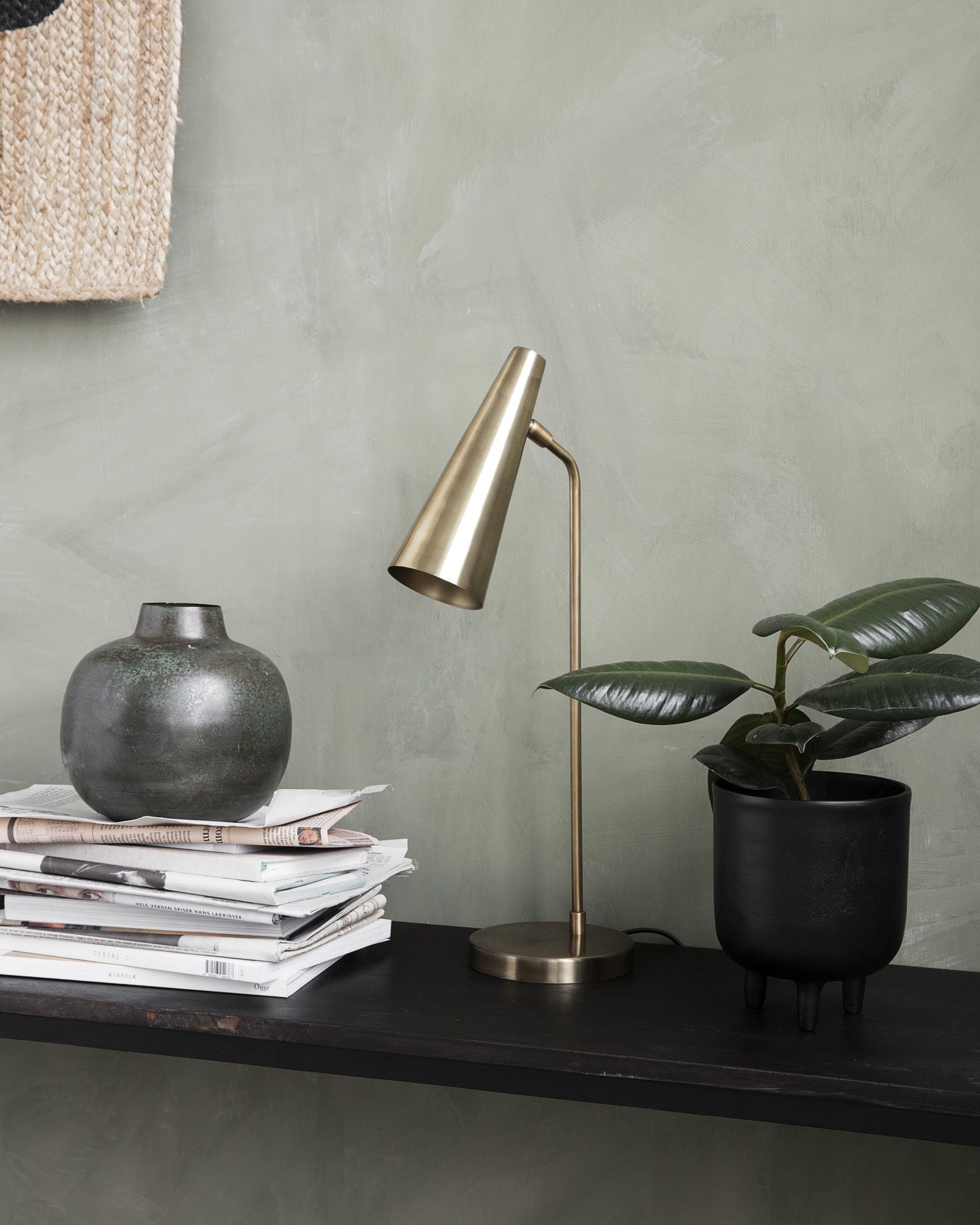 A dark shelf against a green wall holds a stack of magazines with a vase, a brass lamp, and a potted rubber plant.