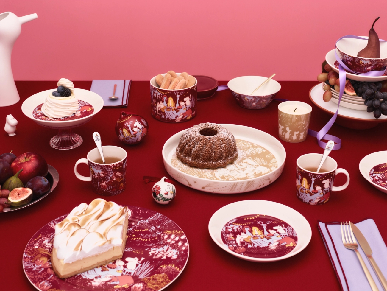 A festive table on a red tablecloth laden with cakes, fresh fruit, and dark red patterned tableware, against a pink background.