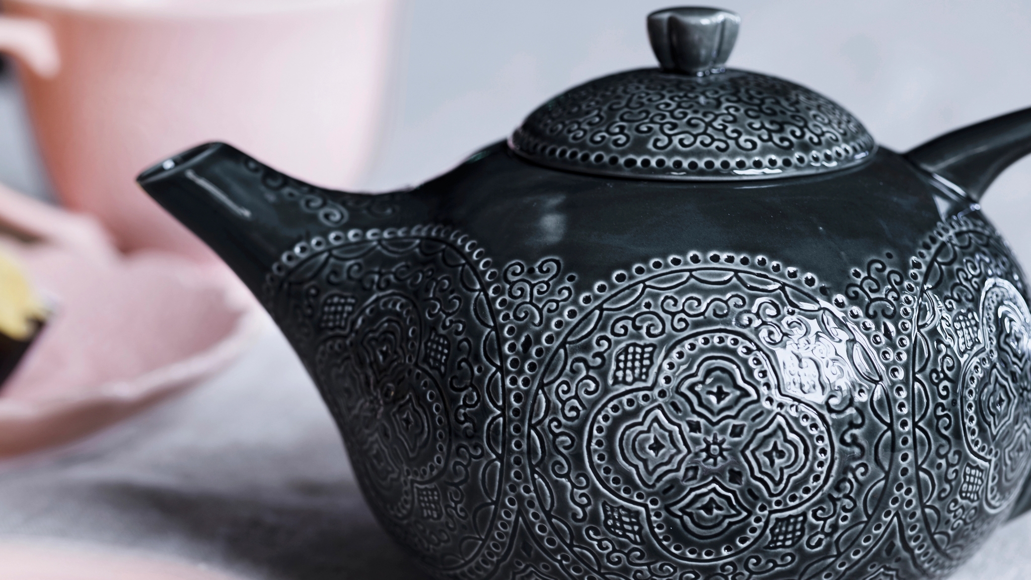 Tummanharmaa, koristeellisilla kaiverruksilla koristeltu teekannu lähikuvassa pöydällä.