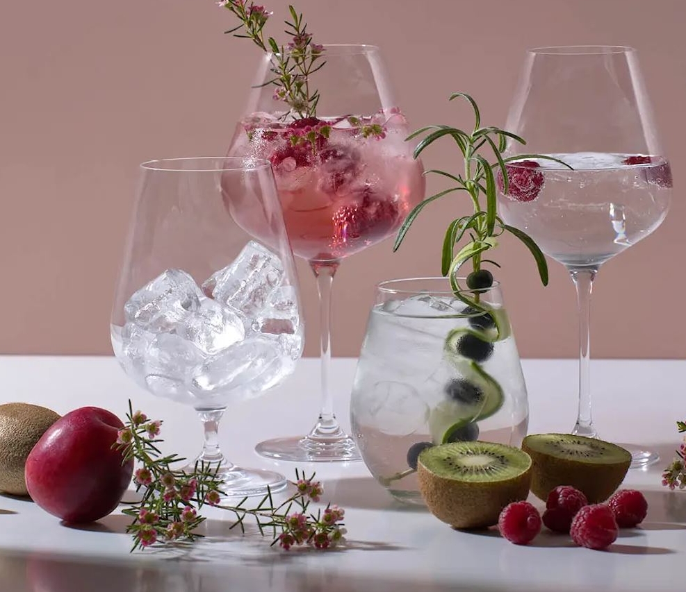 Stilfulla drinkar med is, bär, kiwi och örter uppställda på ett bord mot en rosa bakgrund.