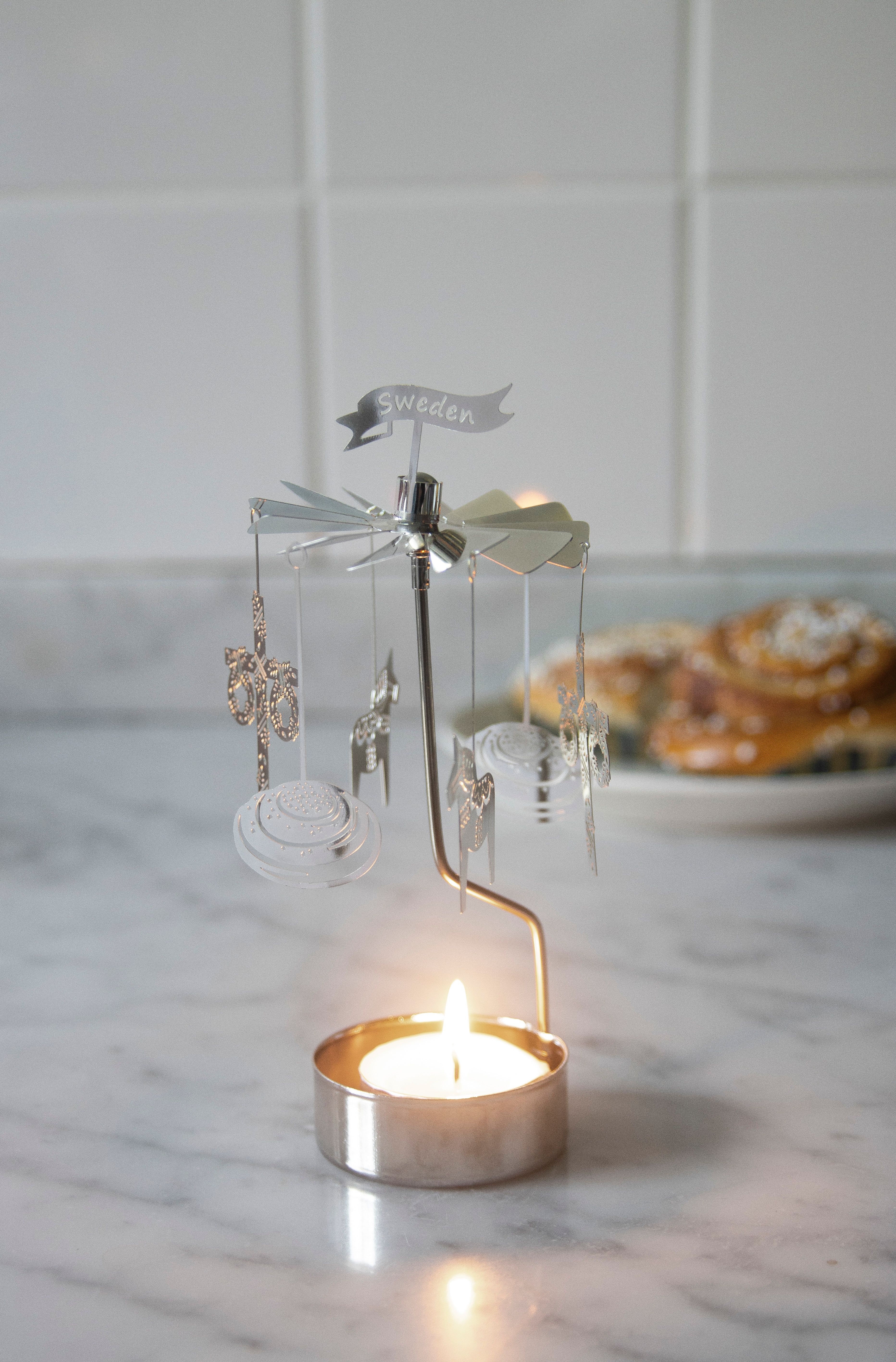 A silver rotating candle holder with a lit tea light, a "Sweden" banner, and dangling charms, with blurred cinnamon rolls in the background.