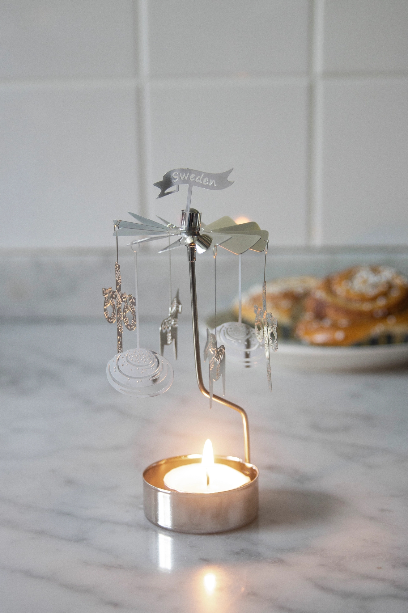 A silver rotating candle holder with a lit tea light, a "Sweden" banner, and dangling charms, with blurred cinnamon rolls in the background.