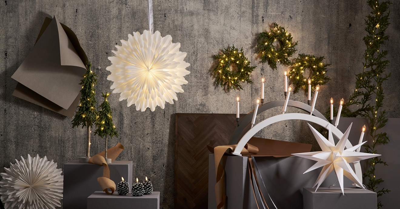 Juhlava kokonaisuus valkoisia paperitähtivaloja, valkoinen kynttelikkö palavine kynttilöineen, minikuusia sekä havukransseja harmaata seinää vasten.