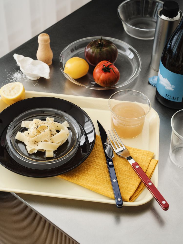 Flat pasta with black pepper, an amber drink, a blue-handled knife, and a red-handled fork on an orange napkin, all on a yellow tray.