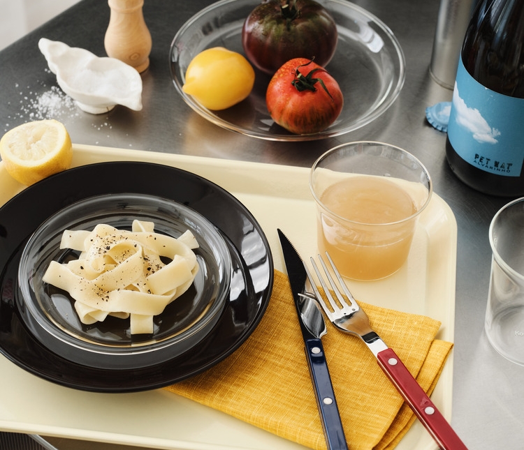 Pasta with pepper, lemon, juice, and red and blue cutlery on a beige tray.
