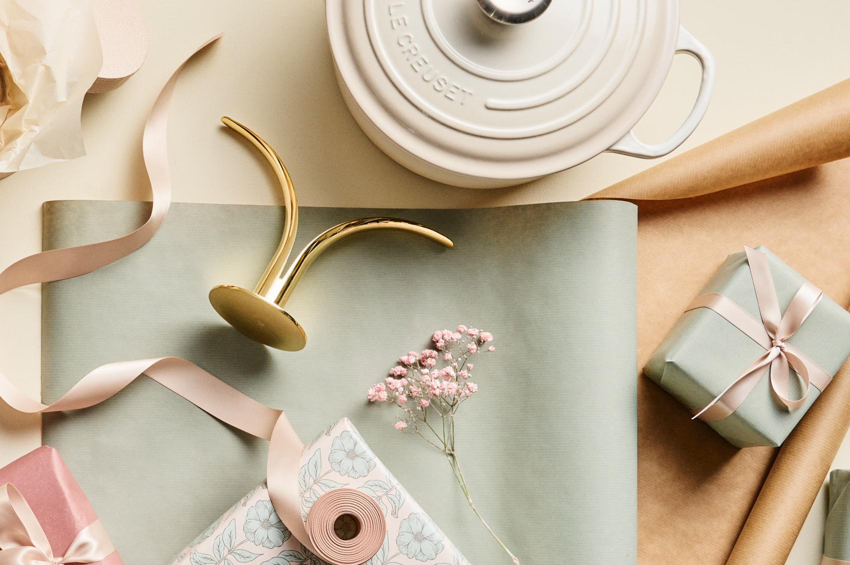 Lahjoja ja paketointitarvikkeita vaalealla taustalla, mukana valkoinen Le Creuset -pata, morsian- ja sulhanen -figuurit, nauhoja ja kukkia.