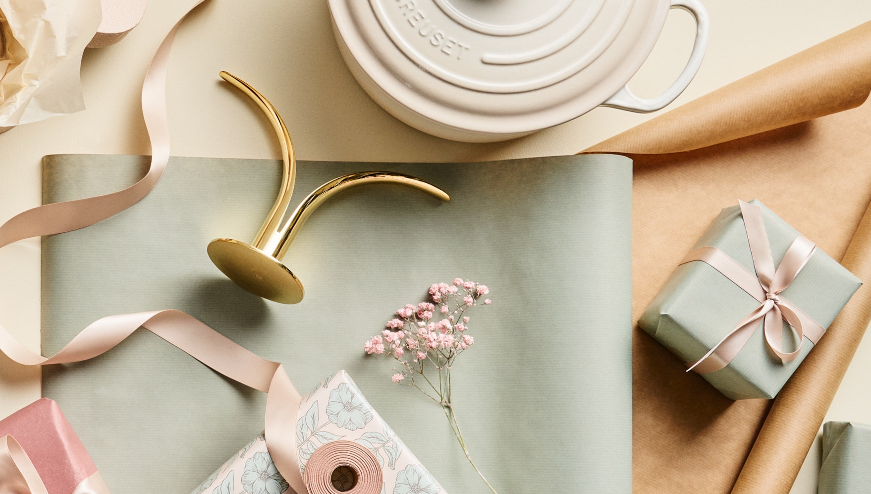 Lahjoja ja paketointitarvikkeita vaalealla taustalla, mukana valkoinen Le Creuset -pata, morsian- ja sulhanen -figuurit, nauhoja ja kukkia.