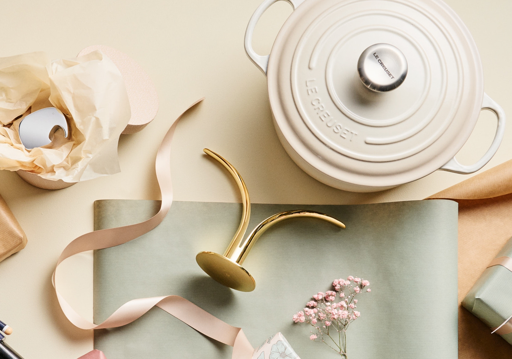 Kermanvärinen Le Creuset -valurautapata, kultainen kynttilänjalka, vaaleanpunainen nauha ja lahjoja aseteltuna vaalealle pinnalle.