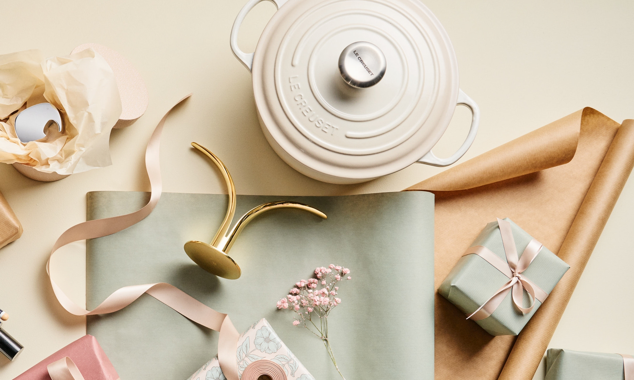 Kermanvärinen Le Creuset -valurautapata, kultainen kynttilänjalka, vaaleanpunainen nauha ja lahjoja aseteltuna vaalealle pinnalle.