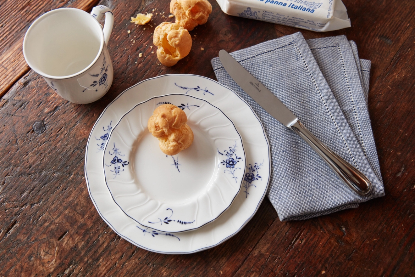 Valkoisia lautasia sinisillä kukkakuvioilla, yhteensopiva muki, useita tuulihattuja, sinisiä lautasliinoja ja veitsi puupinnalla.