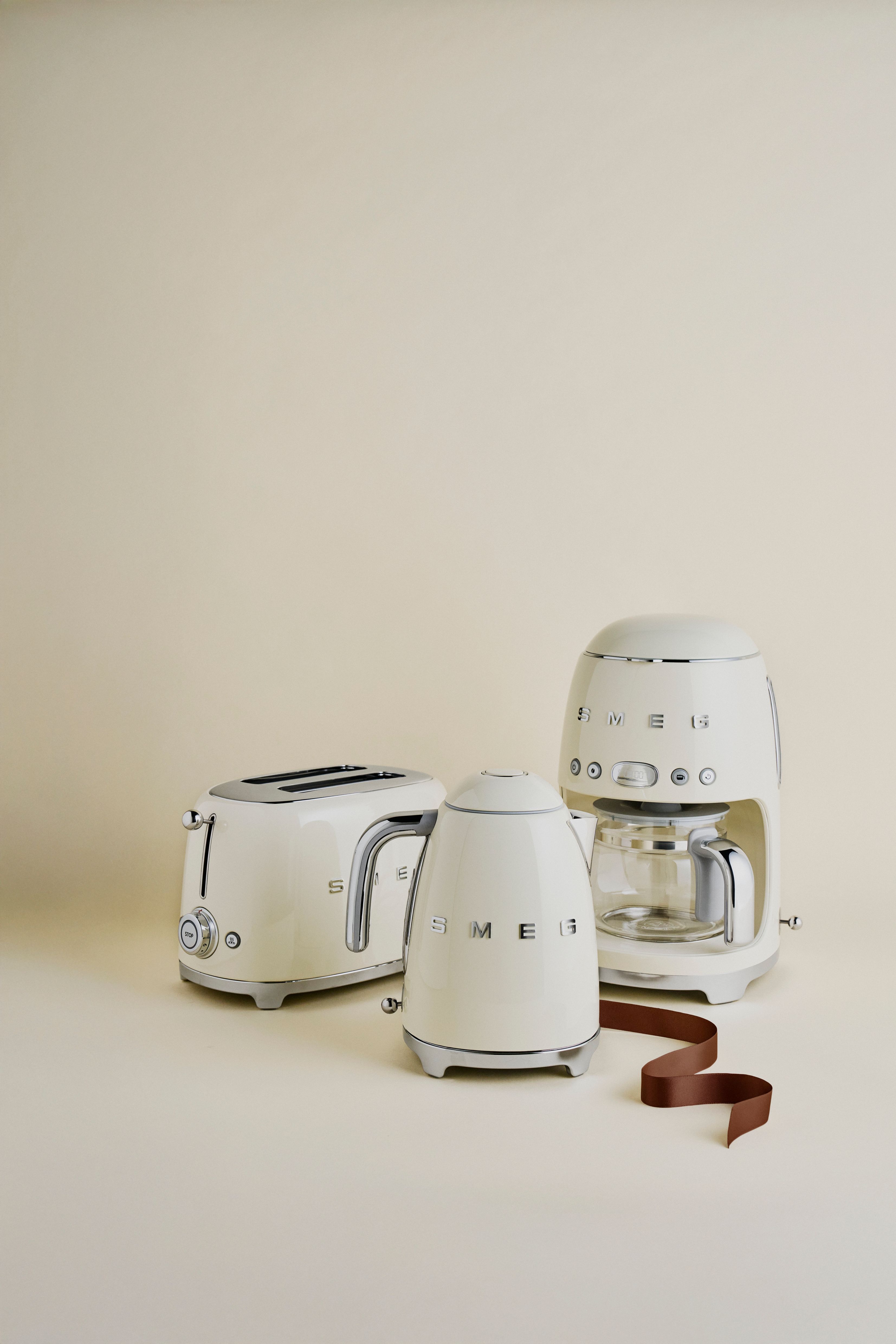 Krämfärgade SMEG köksmaskiner – brödrost, vattenkokare och kaffebryggare – står stilrent arrangerade mot en beige bakgrund.