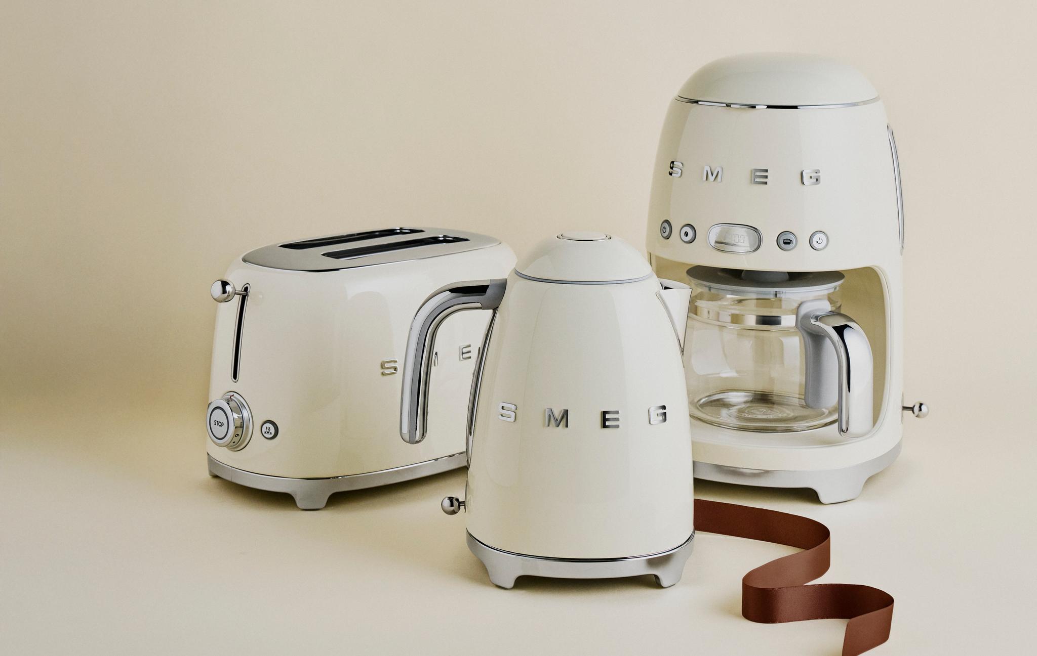 Krämfärgade SMEG köksmaskiner – brödrost, vattenkokare och kaffebryggare – står stilrent arrangerade mot en beige bakgrund.