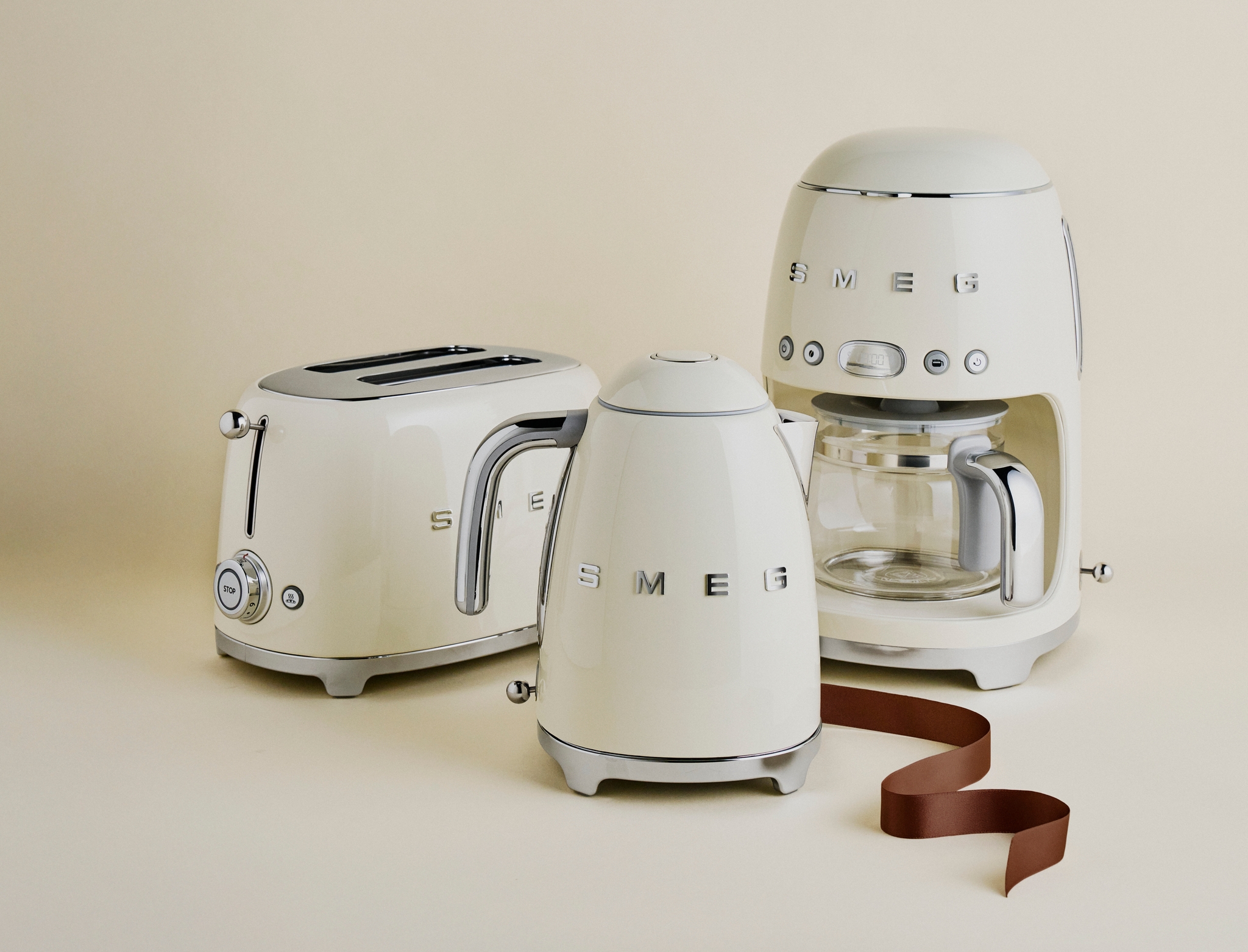 Krämfärgade SMEG köksapparater – brödrost, vattenkokare och kaffebryggare – står uppställda mot en beige bakgrund.