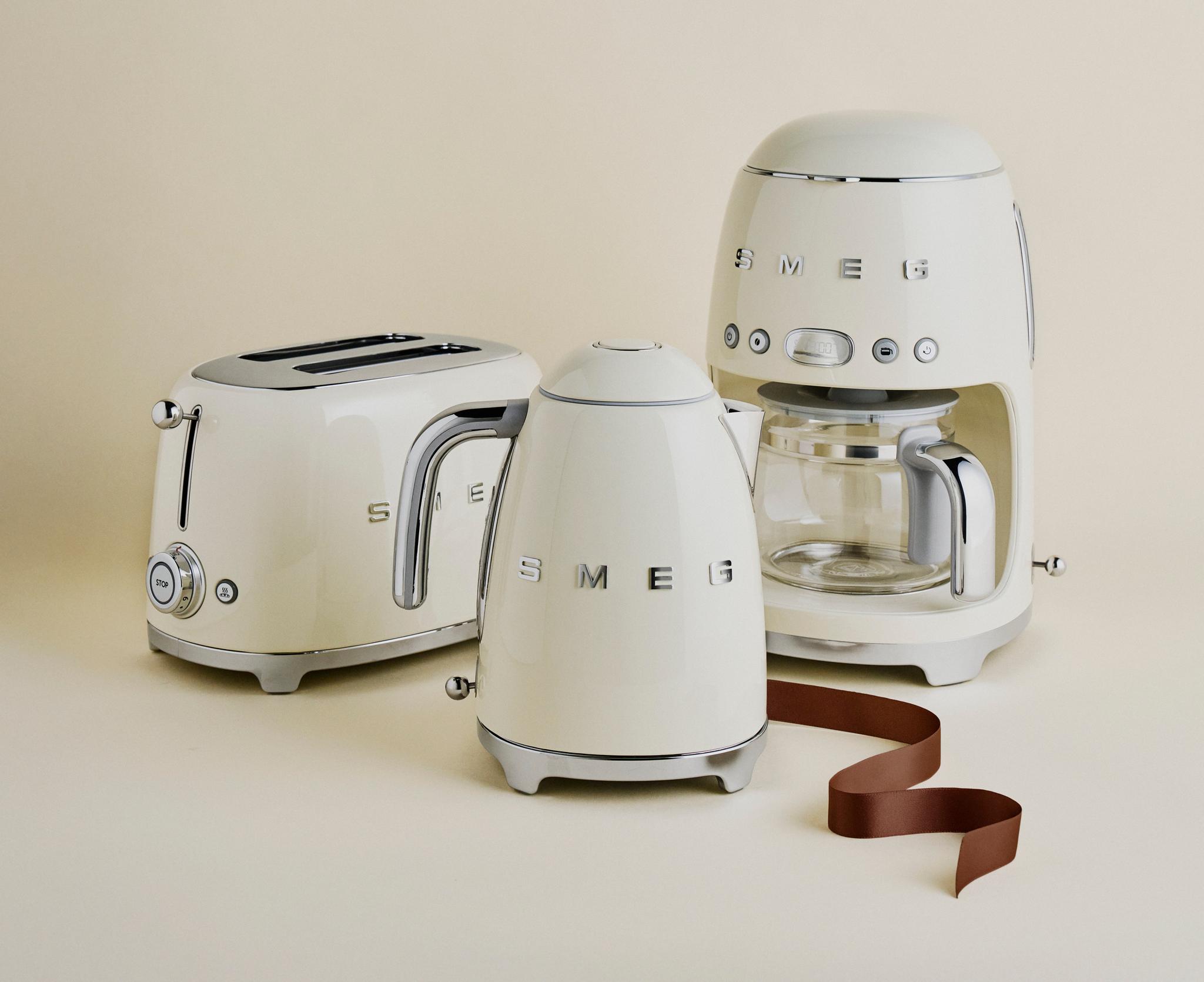 Krämfärgade SMEG köksmaskiner – brödrost, vattenkokare och kaffebryggare – står uppställda mot en beige bakgrund.