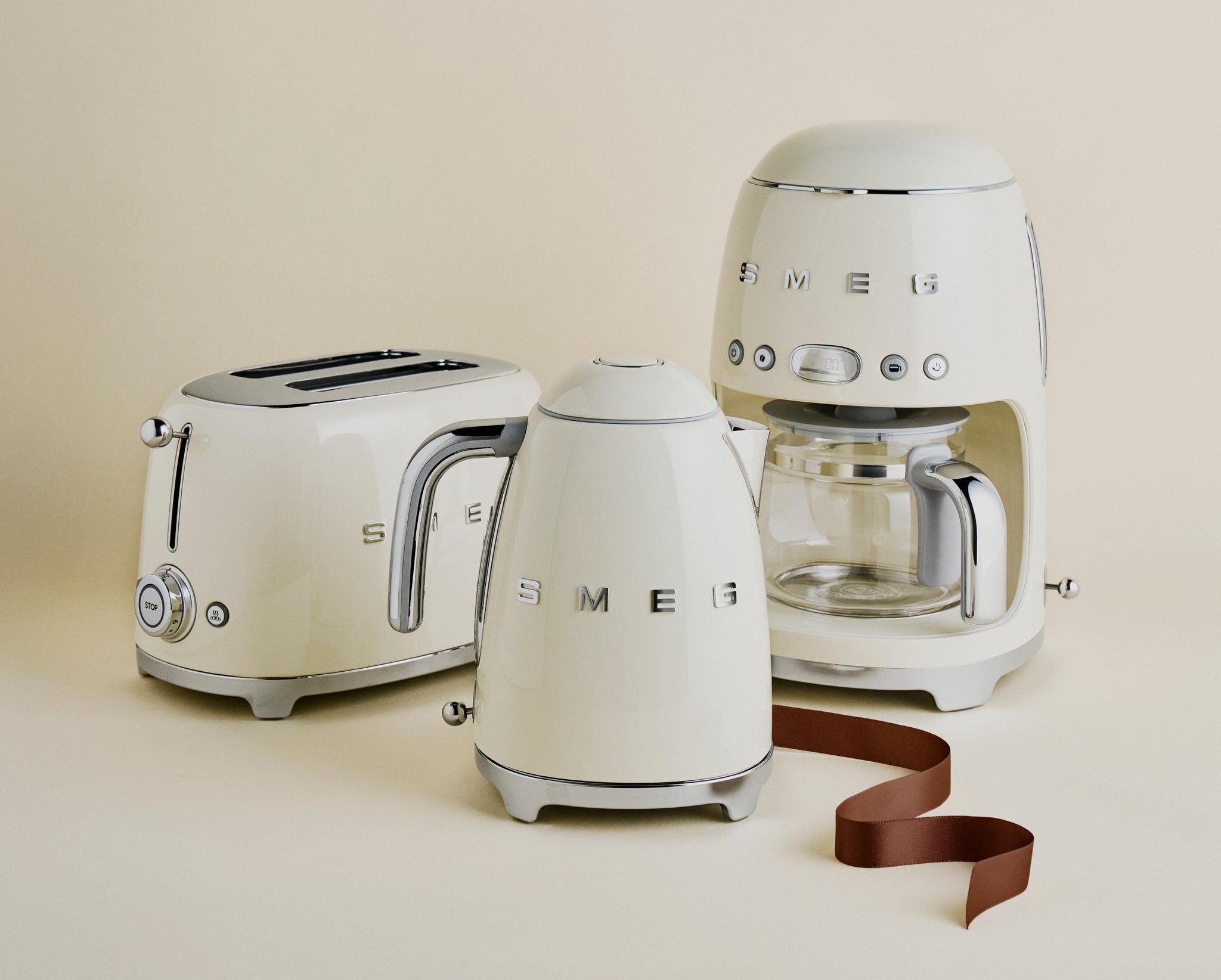 Kremfargede SMEG kjøkkenapparater – brødrister, vannkoker og kaffetrakter – står stilrent oppstilt mot en lys beige bakgrunn.
