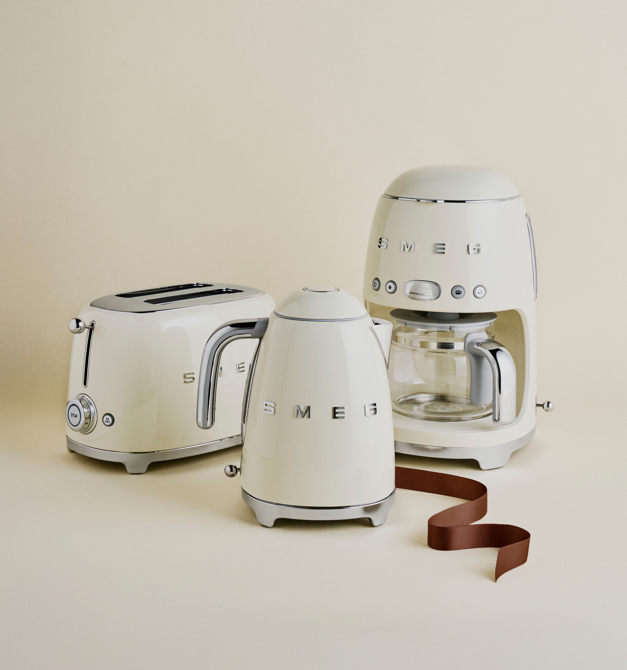 Krämfärgade SMEG köksmaskiner – brödrost, vattenkokare och kaffebryggare – står stilrent arrangerade mot en beige bakgrund.